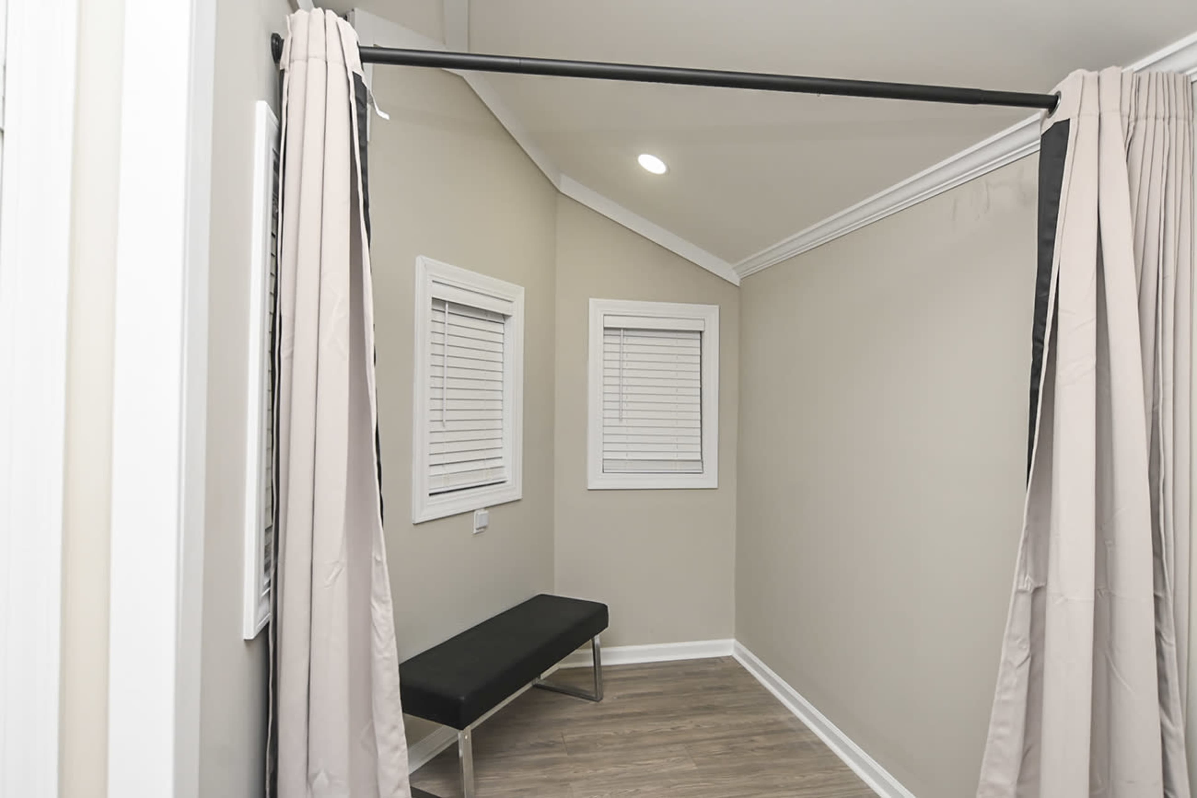 The image shows a small fitting room with beige walls, two windows with white shutters, a black bench, and curtains hung on a rod.