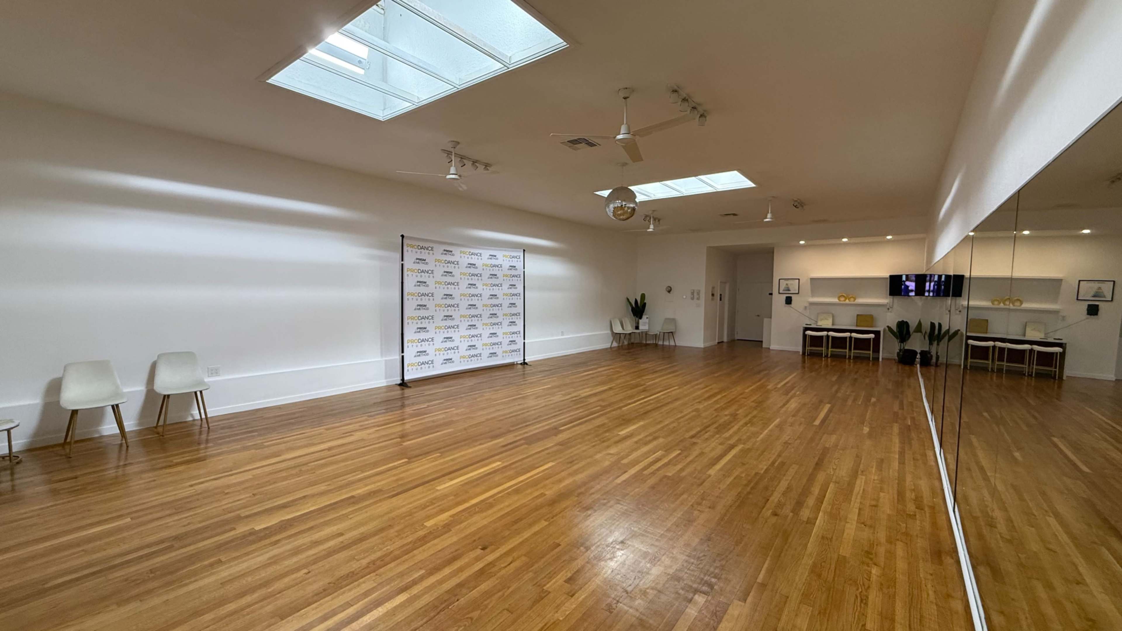 The image shows a spacious, well-lit room with wooden flooring, a large mirror on one wall, and minimalist furniture including chairs and a table.