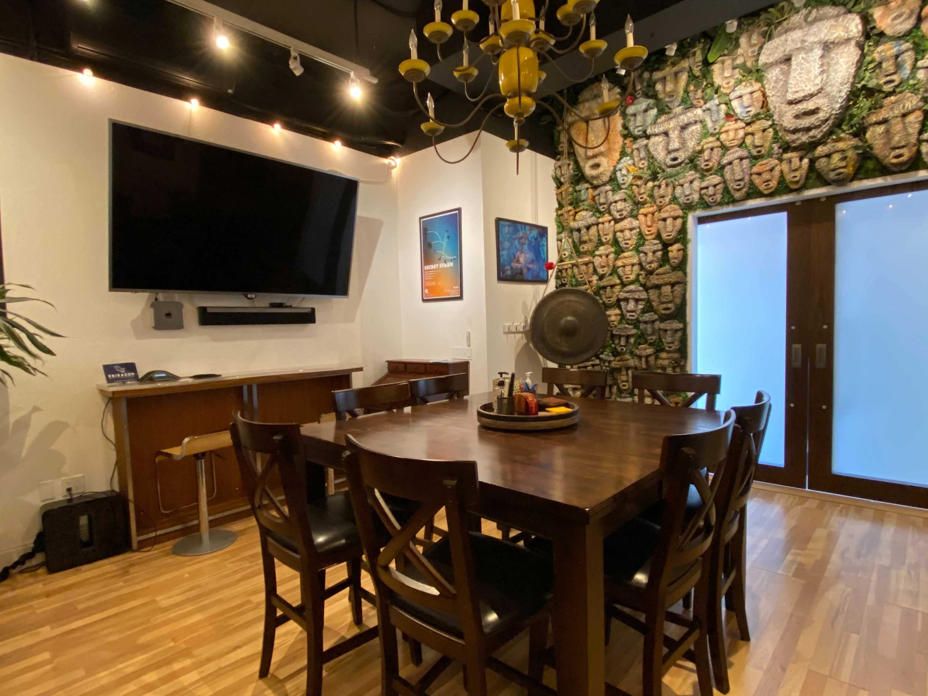 A dining room features a large wooden table surrounded by chairs, a wall decorated with stone face sculptures, and a large television mounted on the wall.