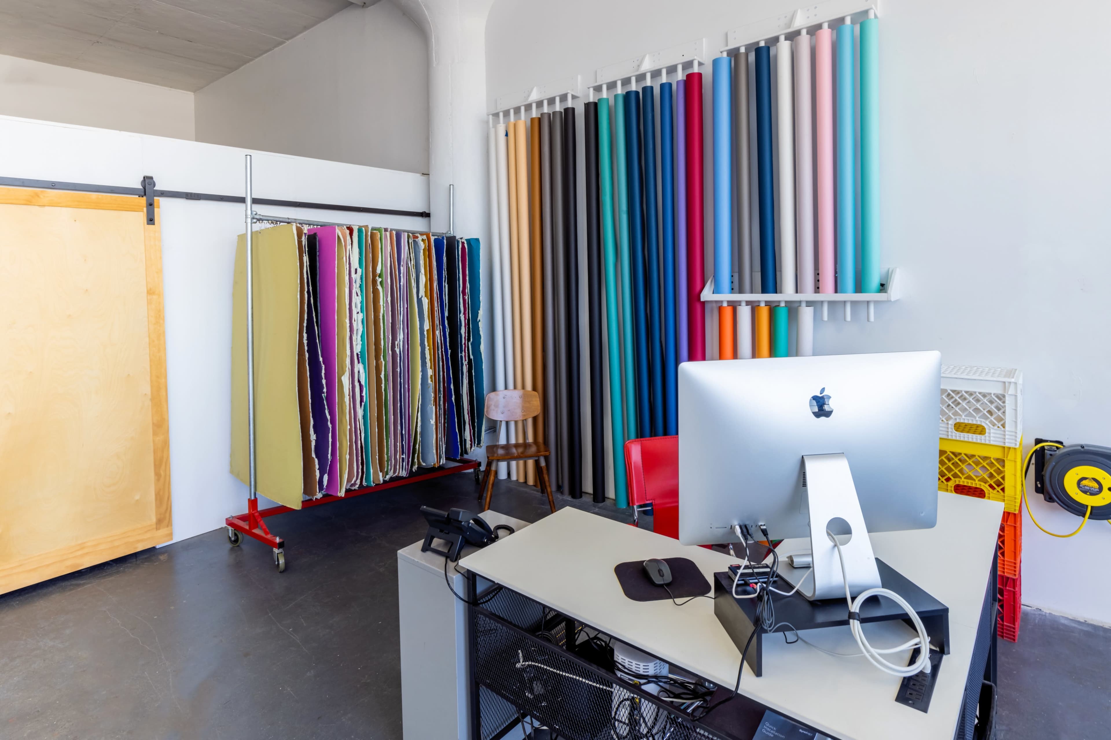 The image shows a workspace with a computer on a desk, alongside a rack of colored paper rolls and a wooden chair in a well-lit room.