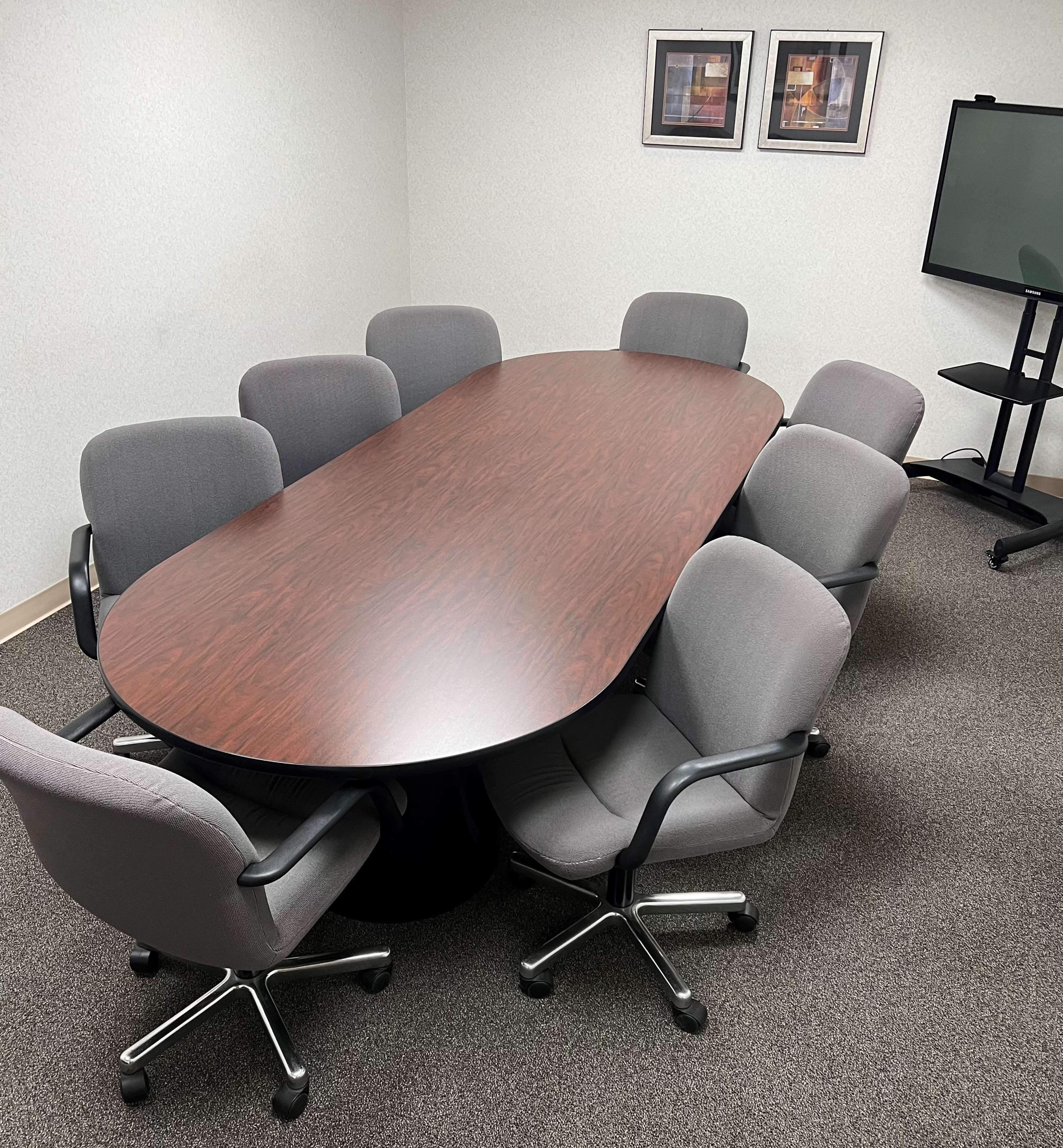 A large oval conference table surrounded by eight gray office chairs is situated in a meeting room.