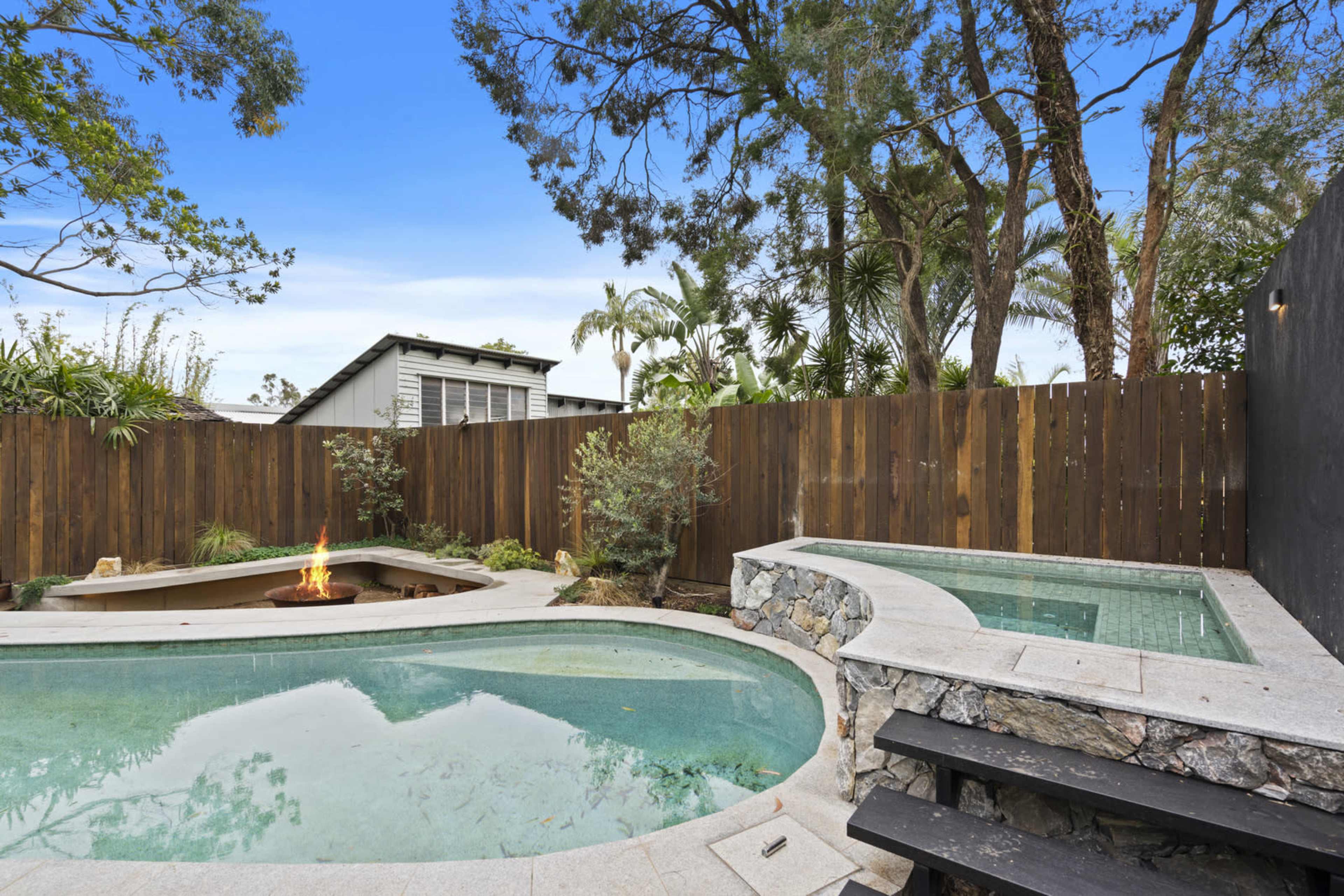 A backyard features a curved swimming pool with a fire pit surrounded by greenery and a wooden fence.