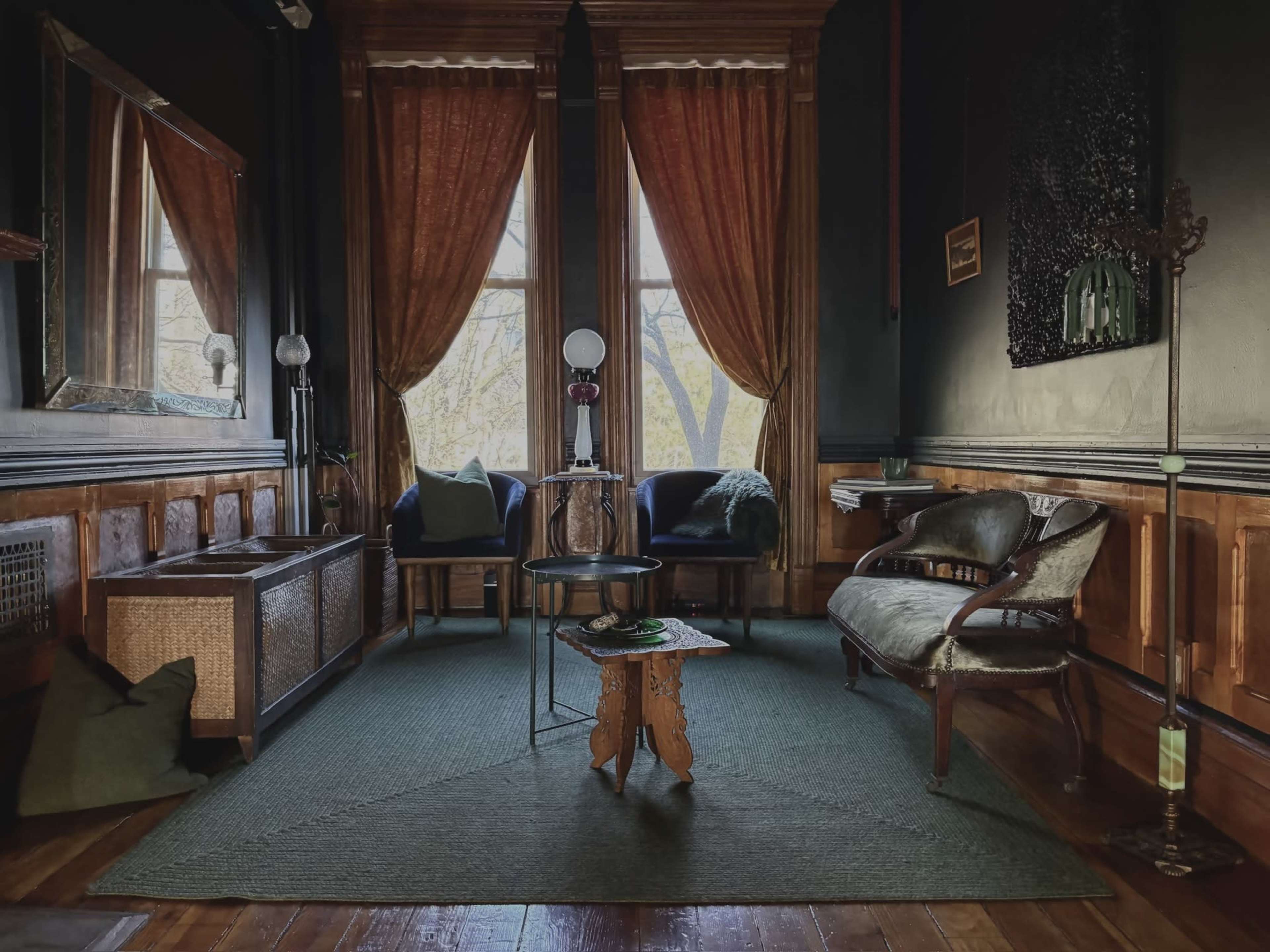 The image shows a cozy parlor with tall windows dressed in orange drapes, featuring a mix of seating, a coffee table, and wooden paneling.