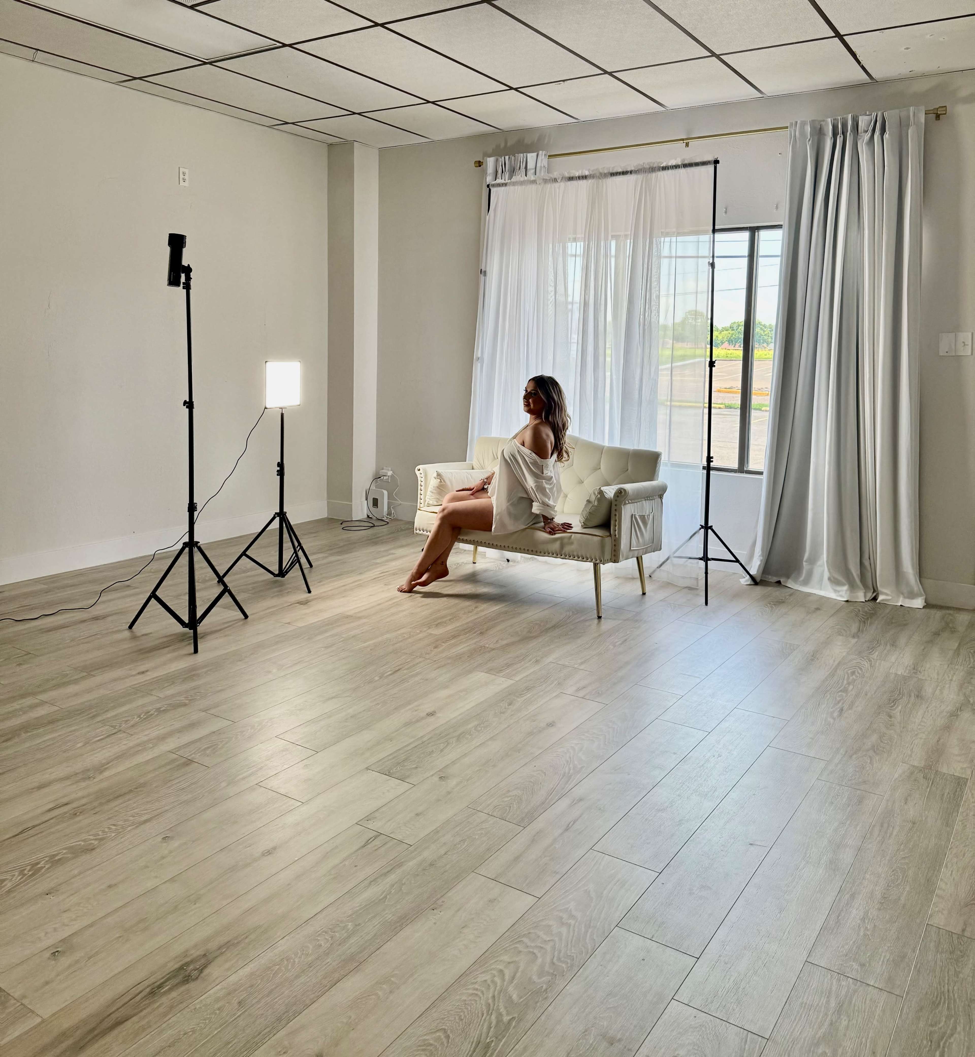 A person sits on a white sofa in a bright, sparsely furnished room with large windows and soft curtains.