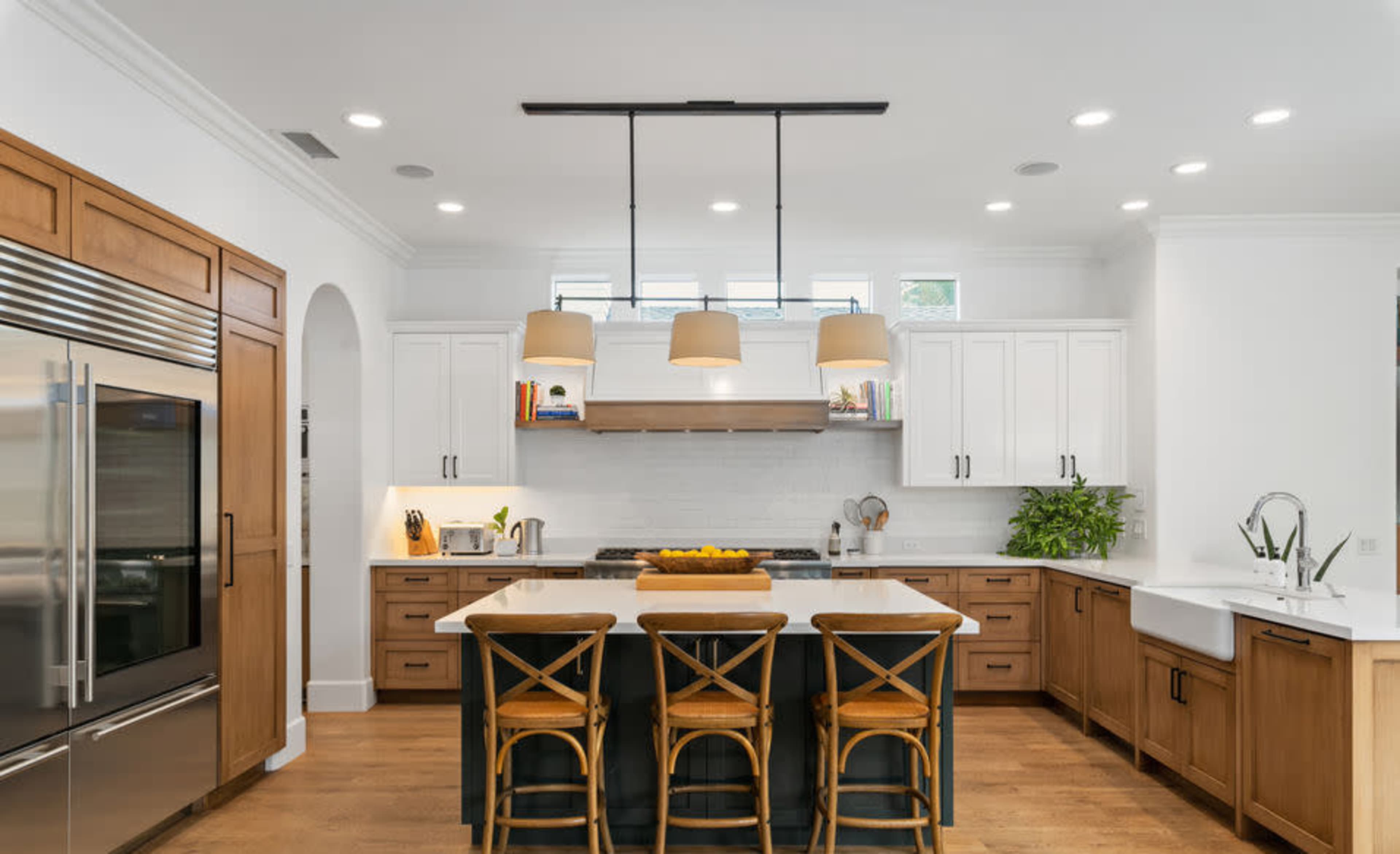 The kitchen features a central island with four wooden stools, surrounded by white cabinets, stainless steel appliances, and pendant lighting.