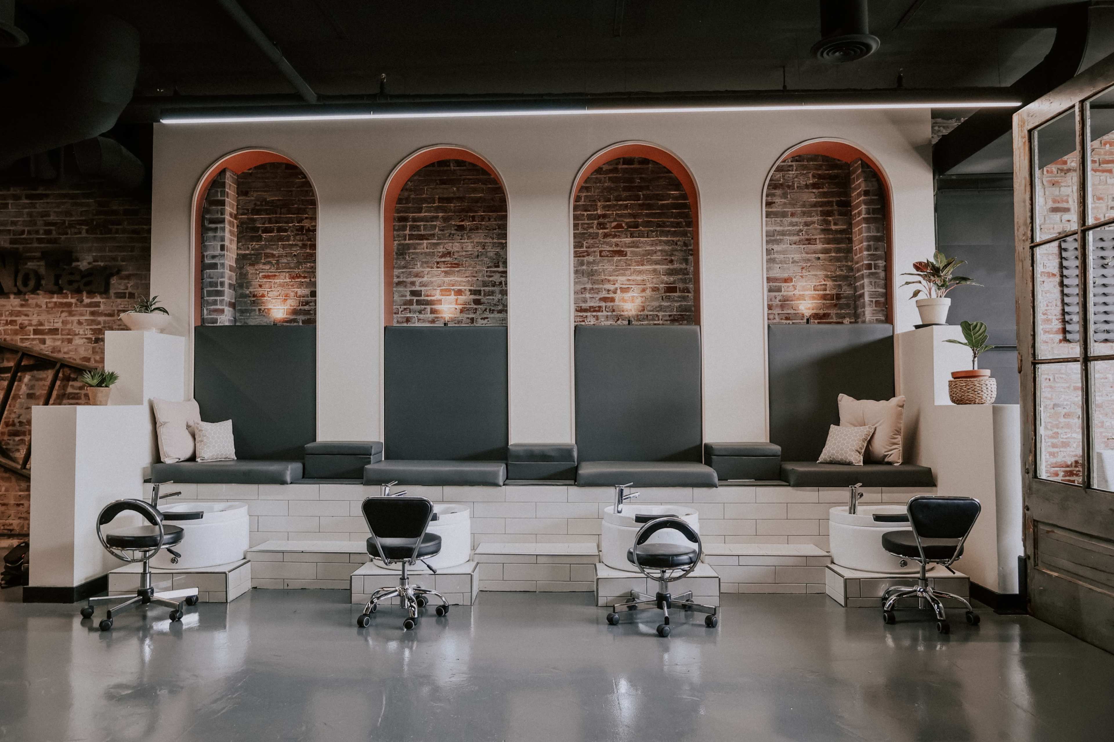 A modern salon interior featuring a series of arched alcoves with seating and pedicure stations, accented by brick walls and minimalist decor.