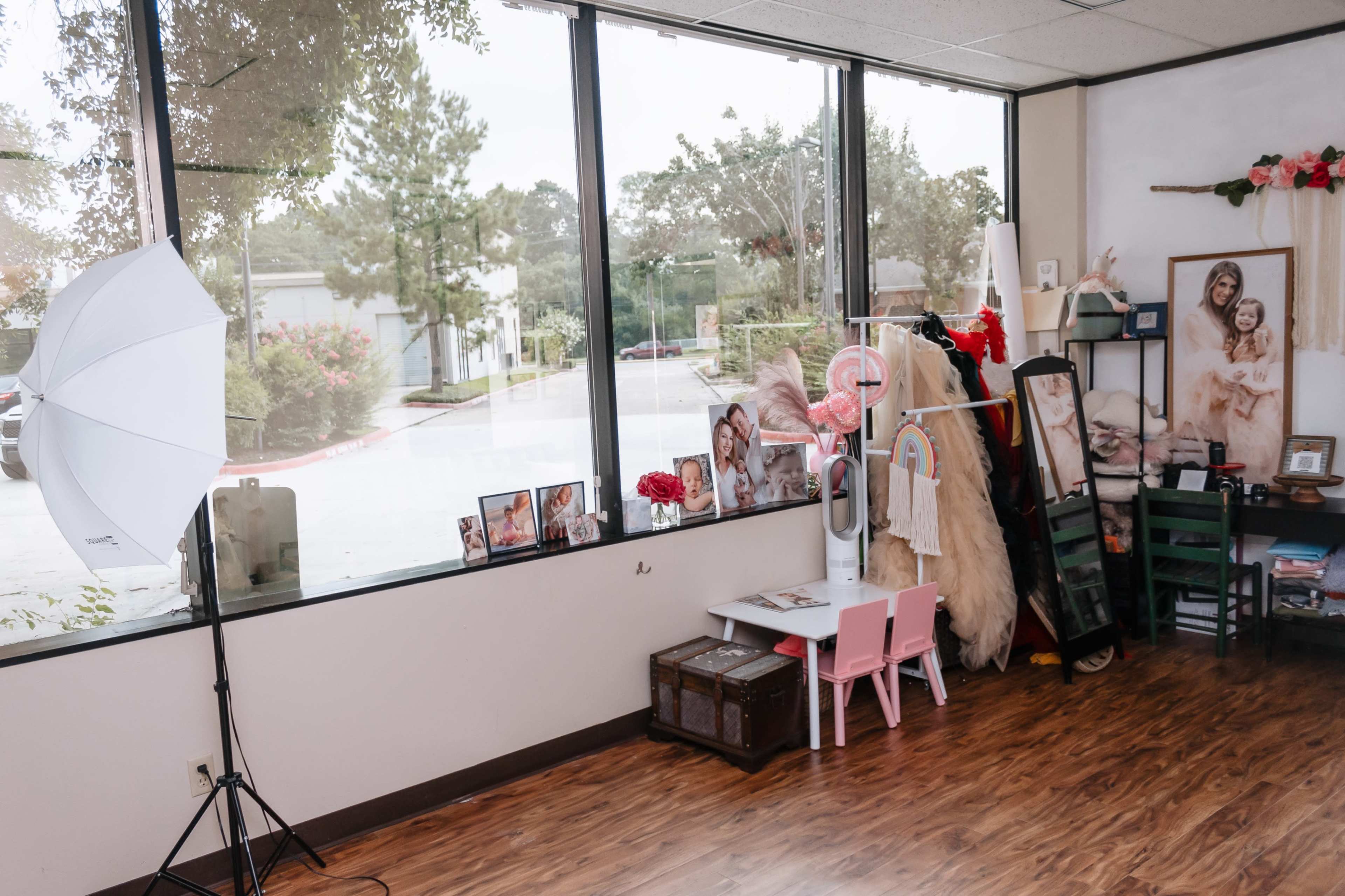 A brightly lit room with large windows, a photography umbrella, a small child-sized table and chairs, and various decorative items, including pictures and clothing, arranged along the walls.