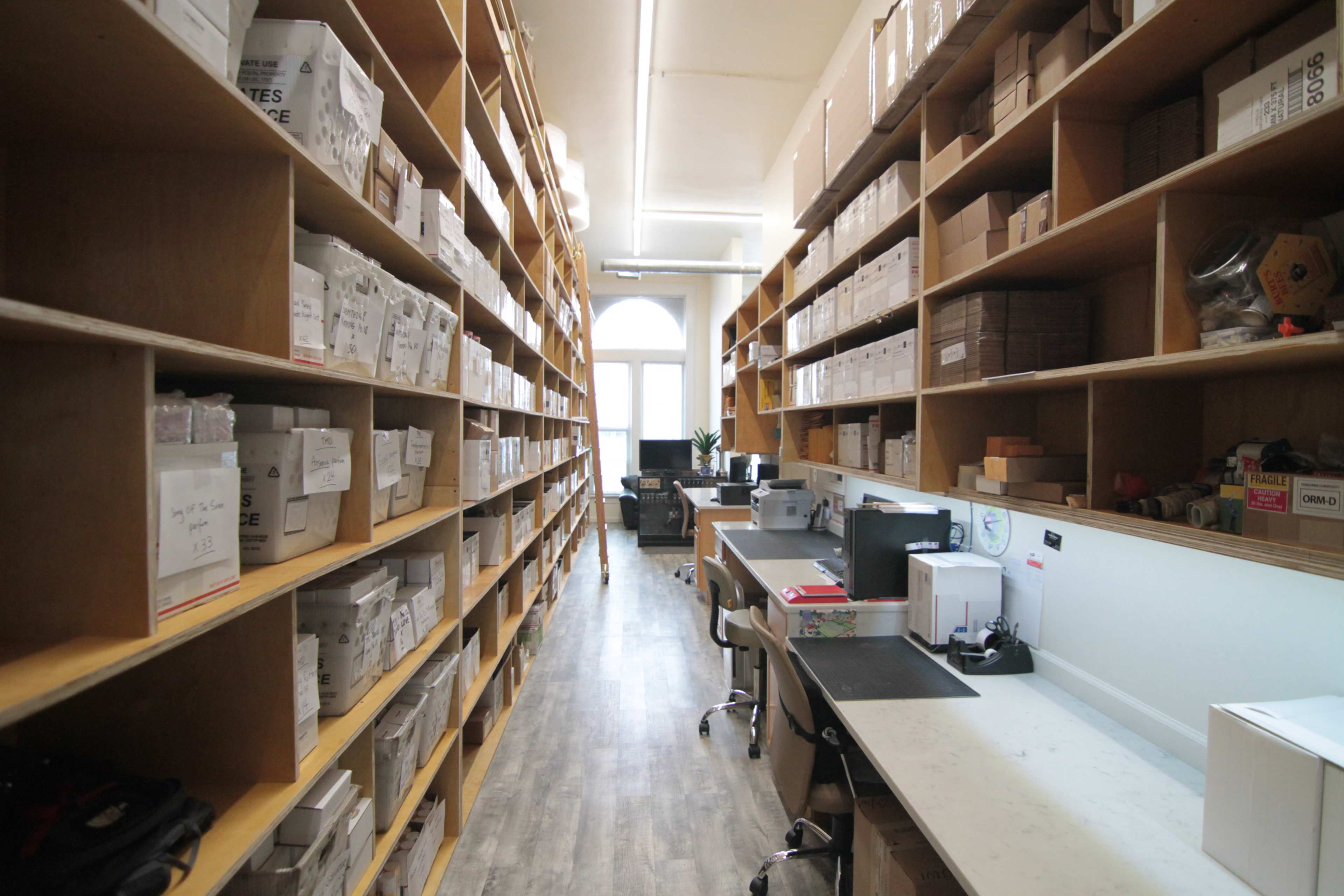 A narrow aisle lined with tall shelves filled with neatly arranged boxes, leading to a workspace with computers and chairs at the far end.