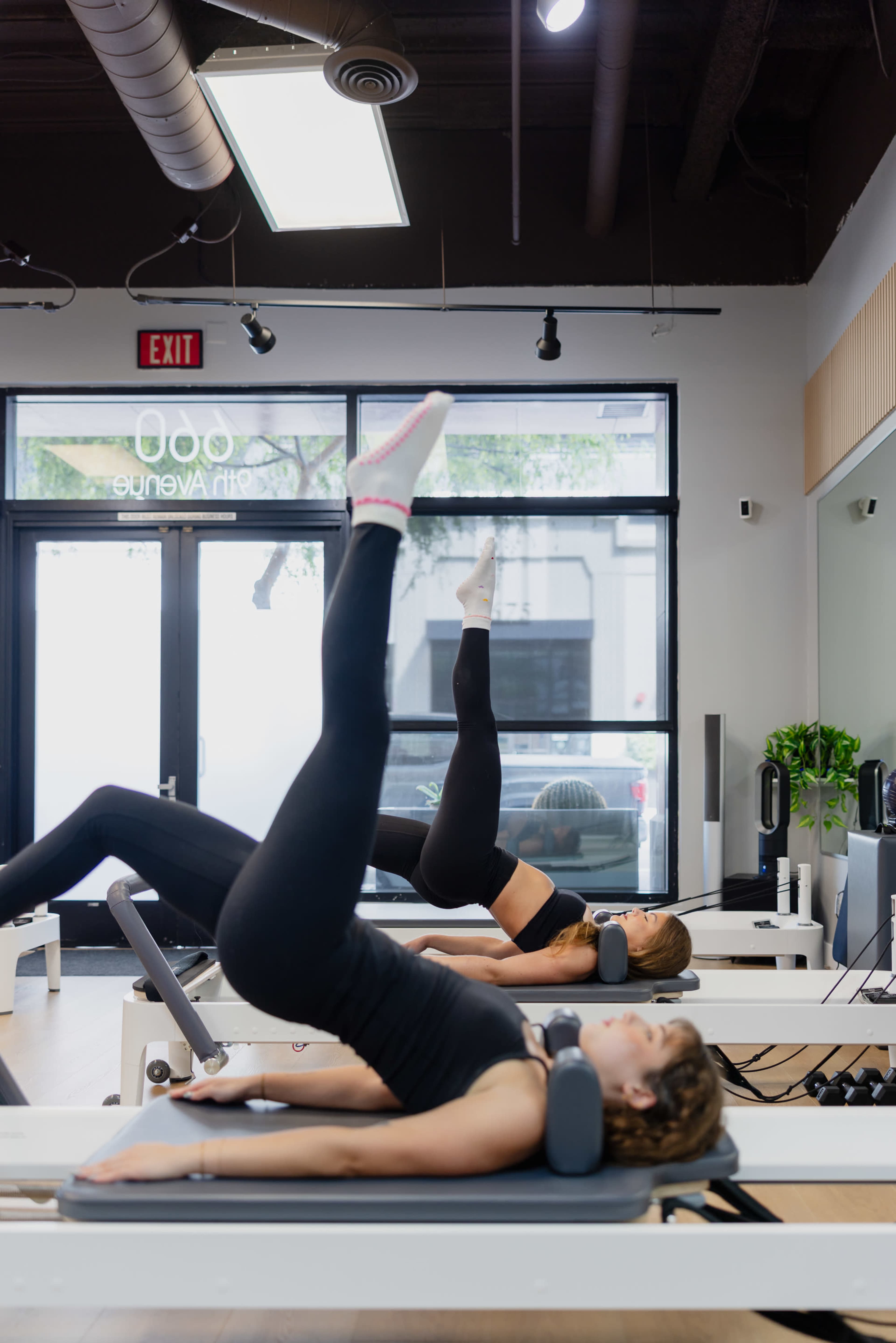 Two women perform leg exercises on reformer Pilates machines in a well-lit studio with large windows.