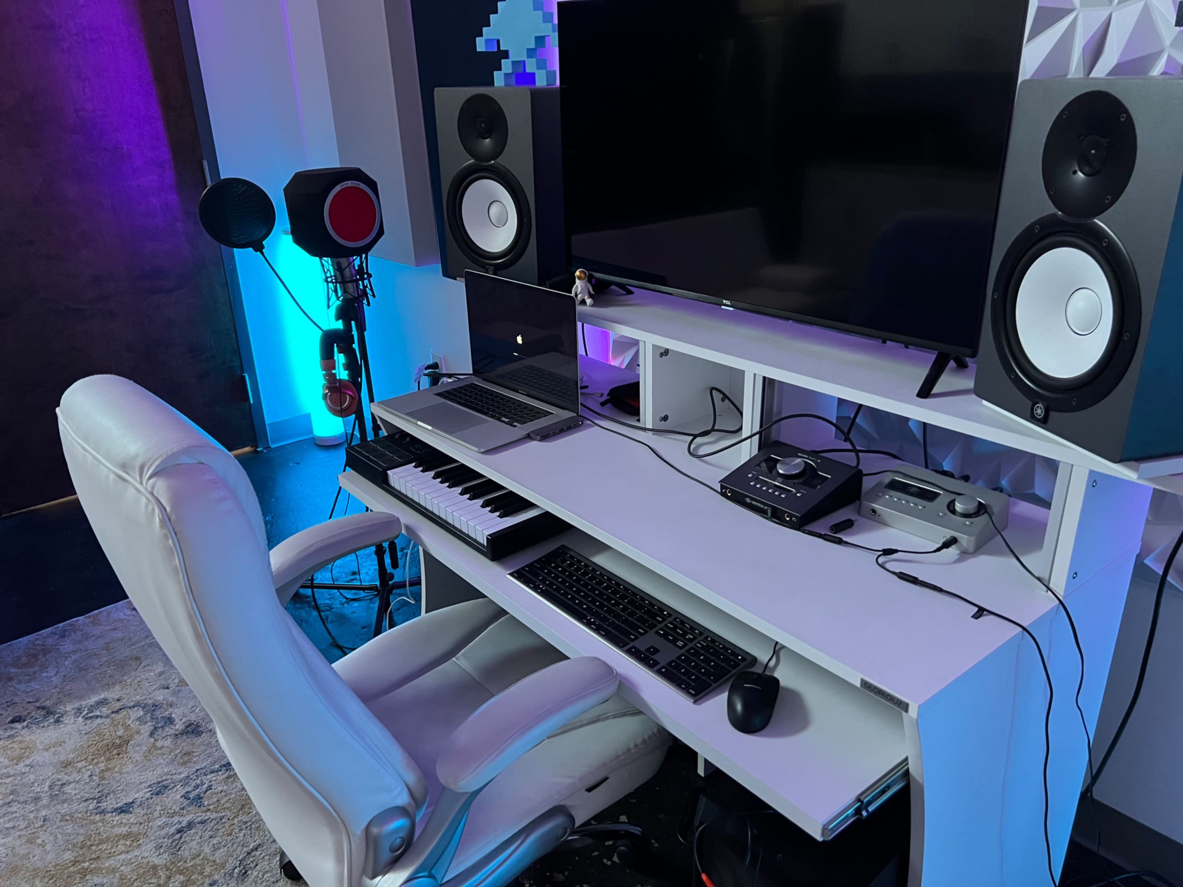 The image shows a modern music production setup featuring a white keyboard, two studio monitors, a computer, and a comfortable chair in a well-lit room.