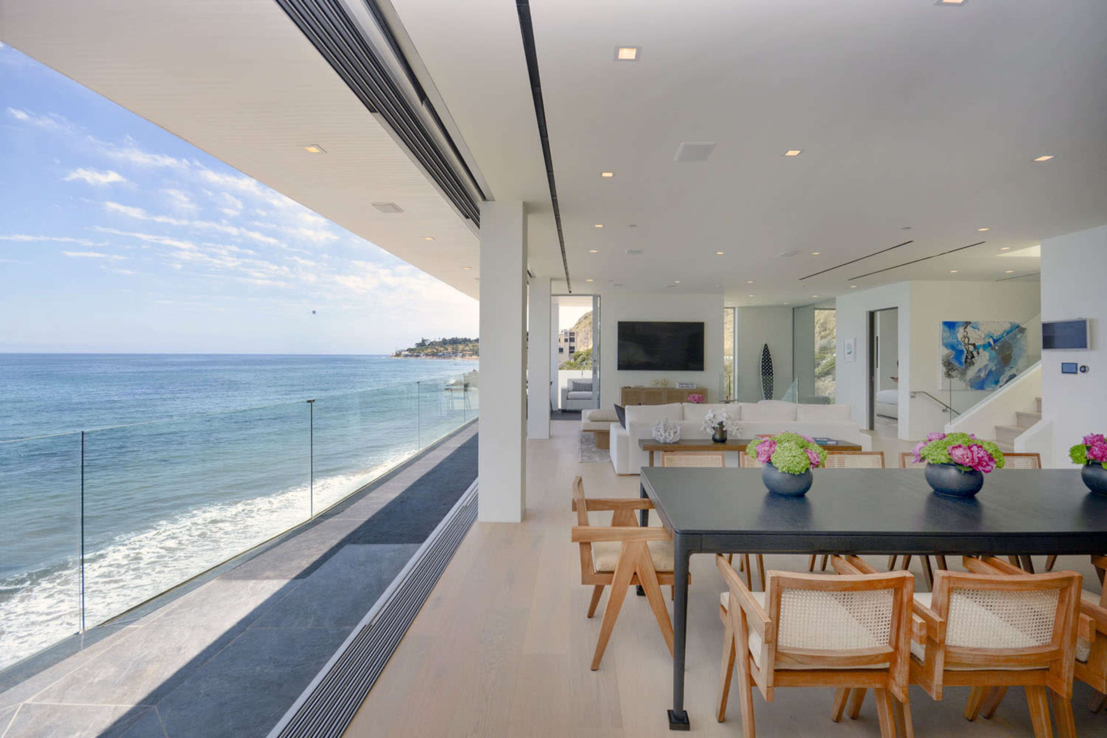 A modern coastal living space features a dining area with a table overlooking a beachfront view through large glass doors.
