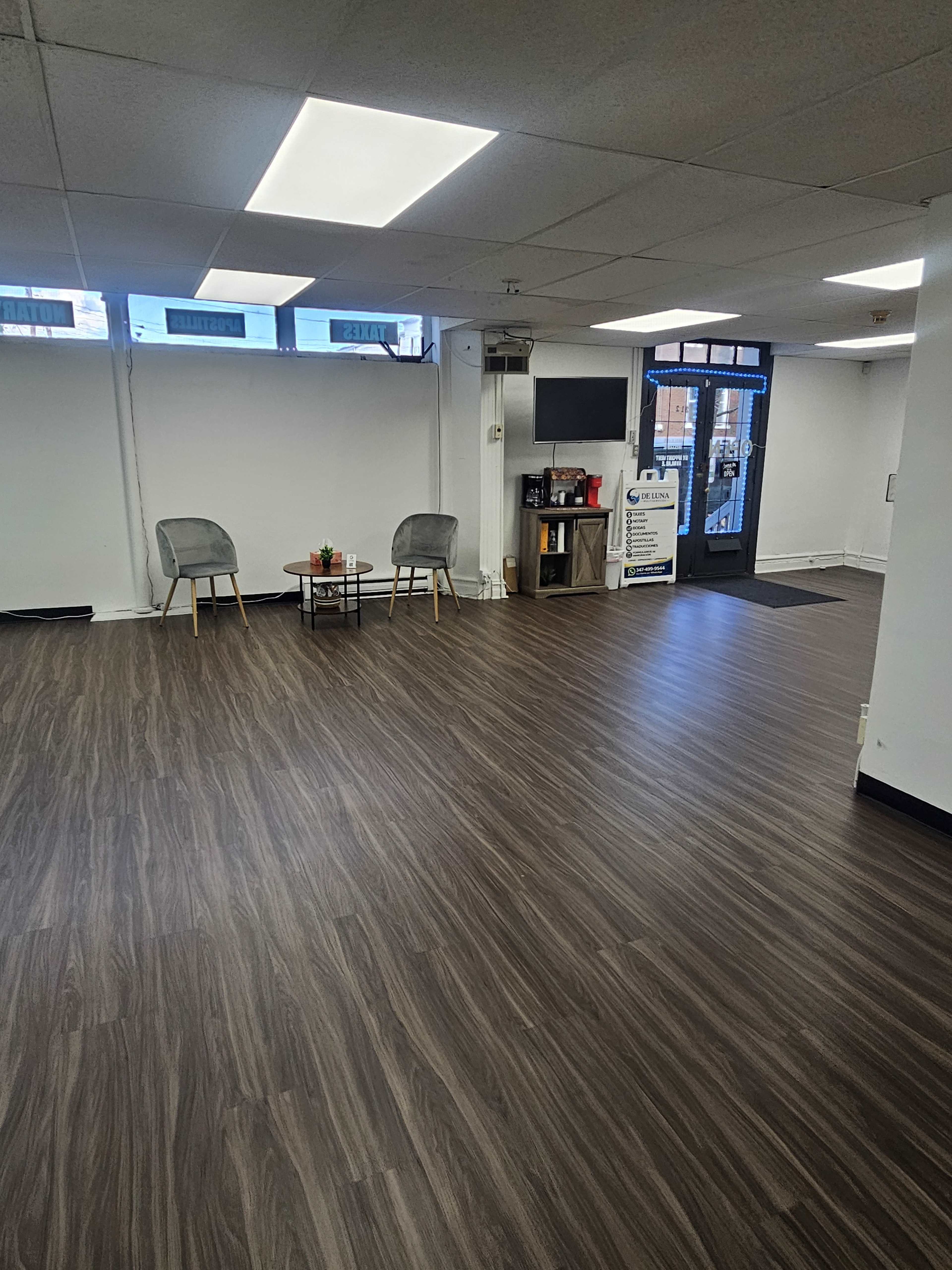 The image shows a spacious, empty room with a wooden floor, two chairs, a small table, and a kitchenette area, along with a doorway leading outside.
