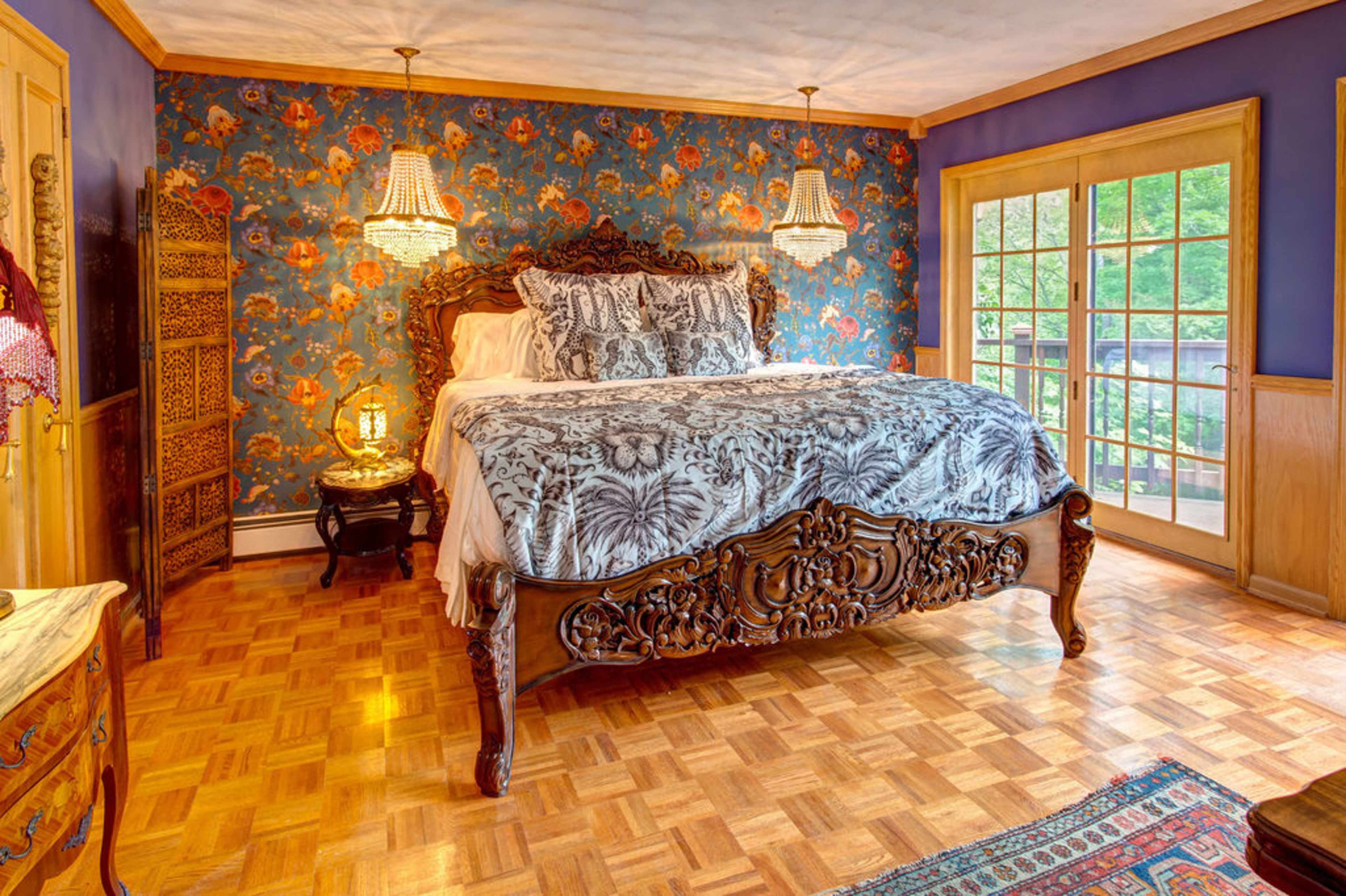 A bedroom features a carved wooden bed with a patterned duvet, ornate light fixtures, and floral wallpaper, complemented by a wooden floor and a view through double doors.