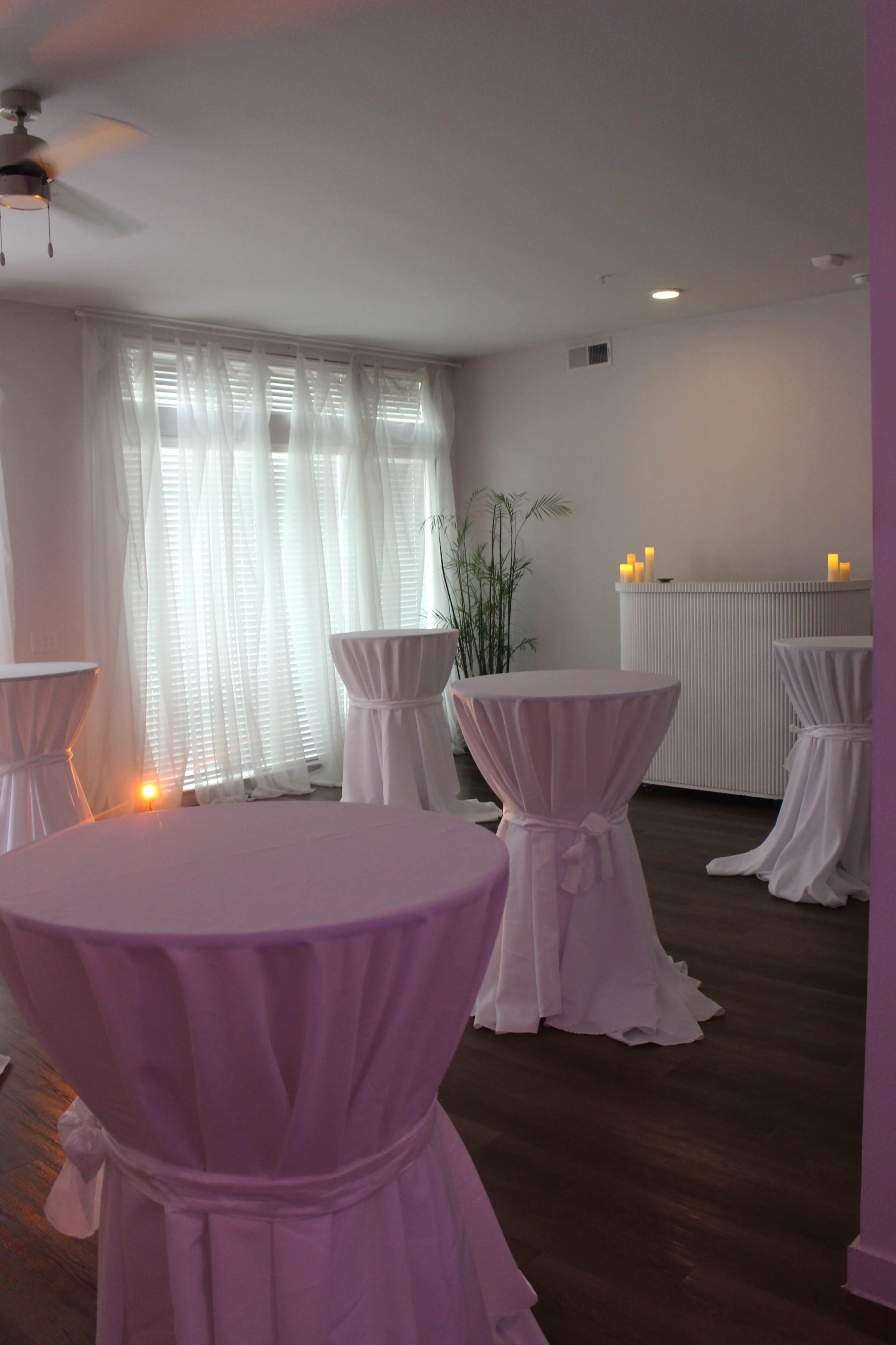 The image shows a well-lit room with several tall tables covered in white cloths, arranged near a window with light drapes and a decorative candle setup along a wall.