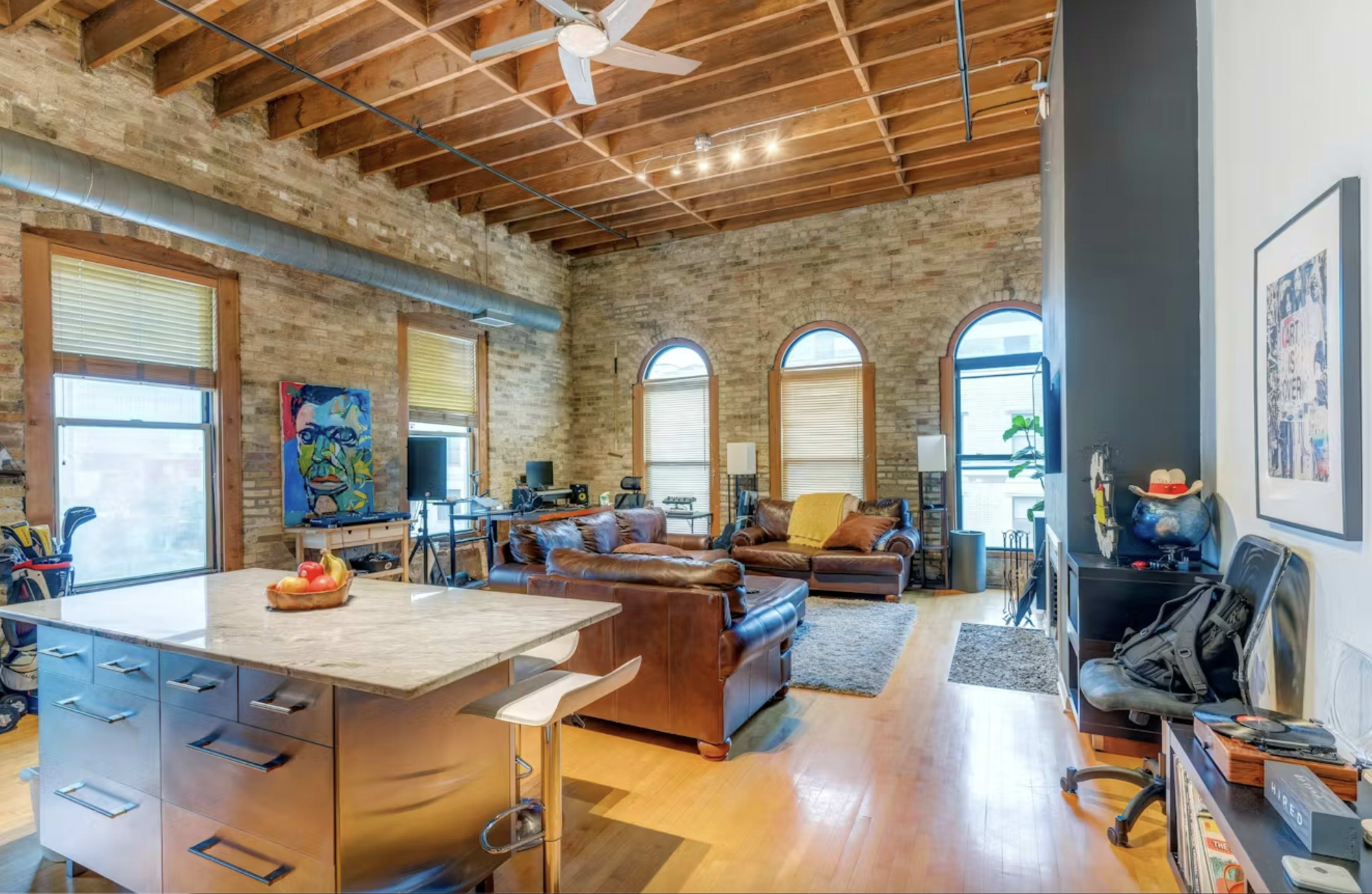 The image shows a spacious loft-style living area featuring exposed brick walls, large windows, and a modern kitchen island.
