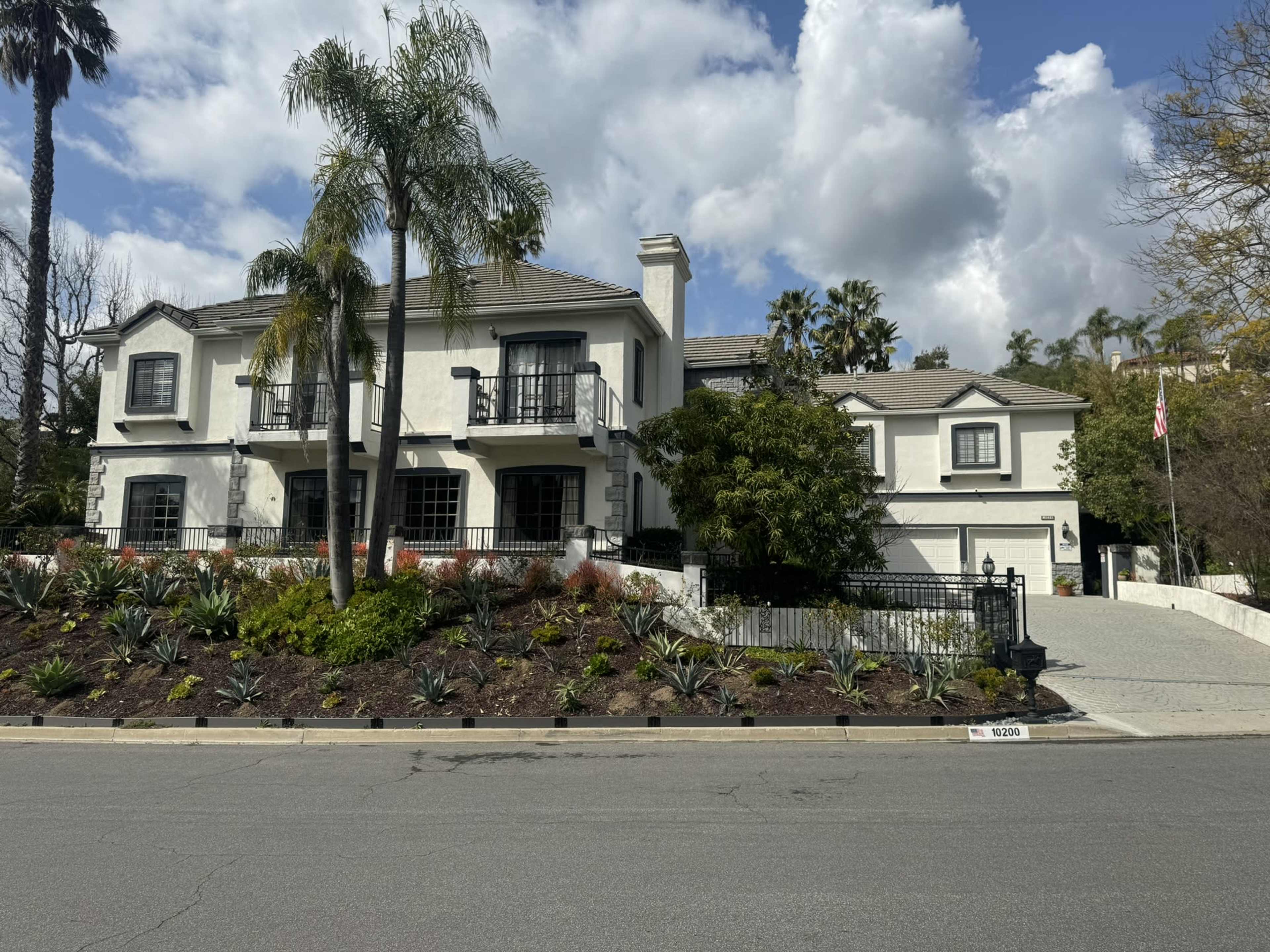 A large, two-story white house with a tiled roof and balcony is set amidst landscaped gardens and palm trees on a suburban street.