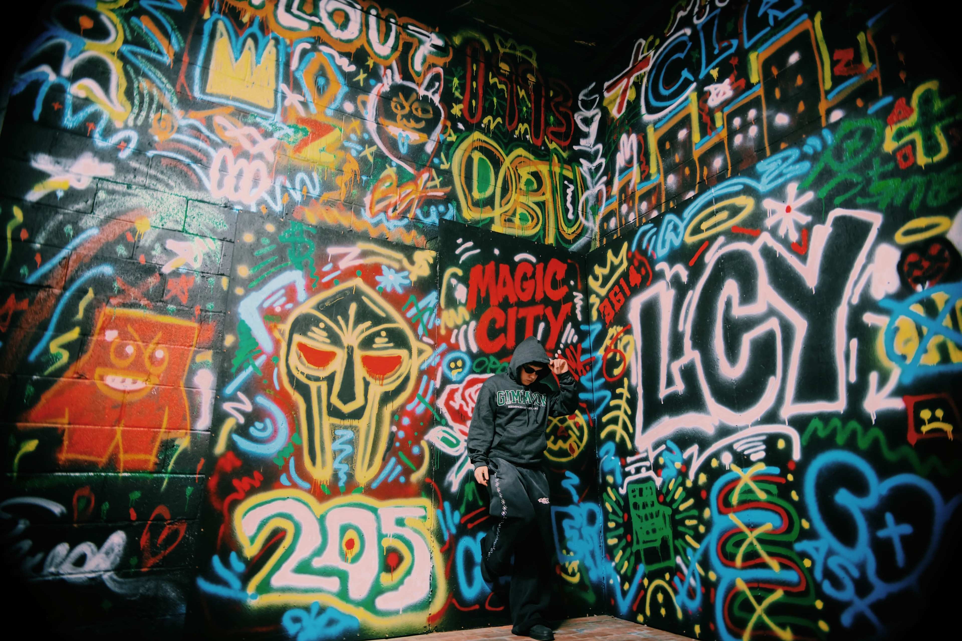 A person stands in front of a colorful graffiti wall covered with various artistic designs and text.