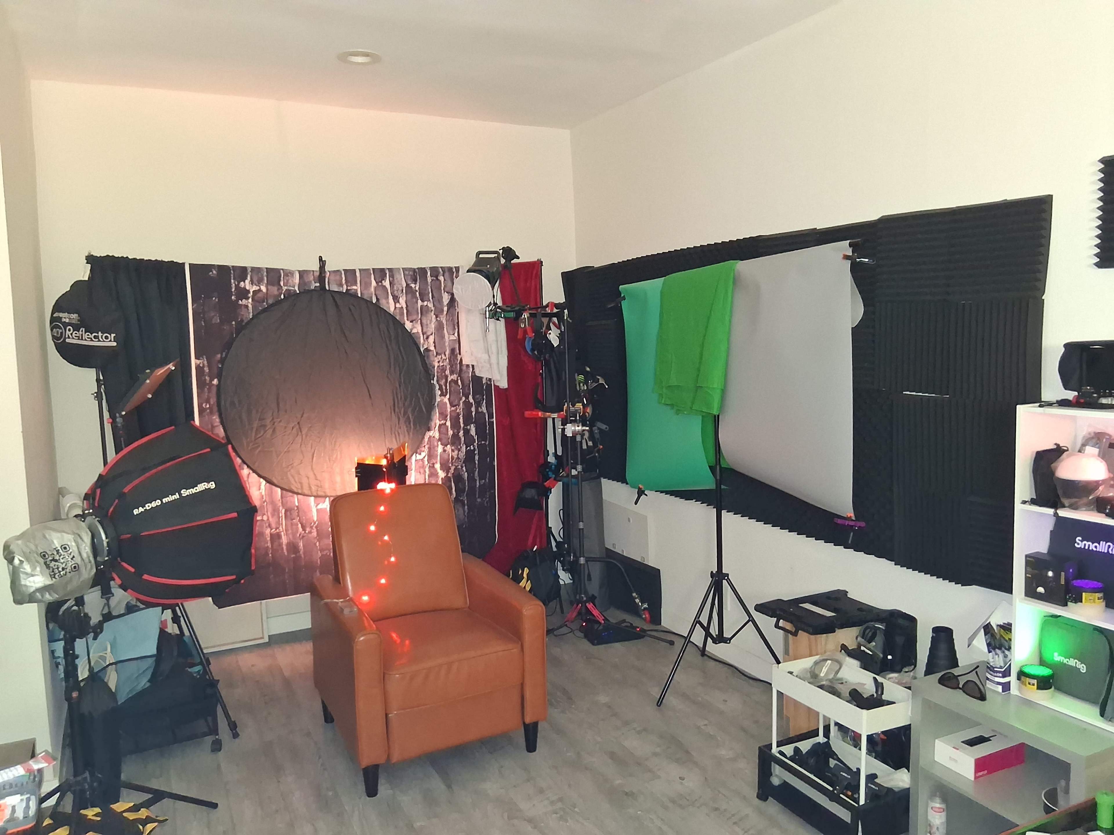 The image shows a photography studio setup featuring a brown chair, various lighting equipment, backdrop stands, and green and gray screens placed against the white walls.