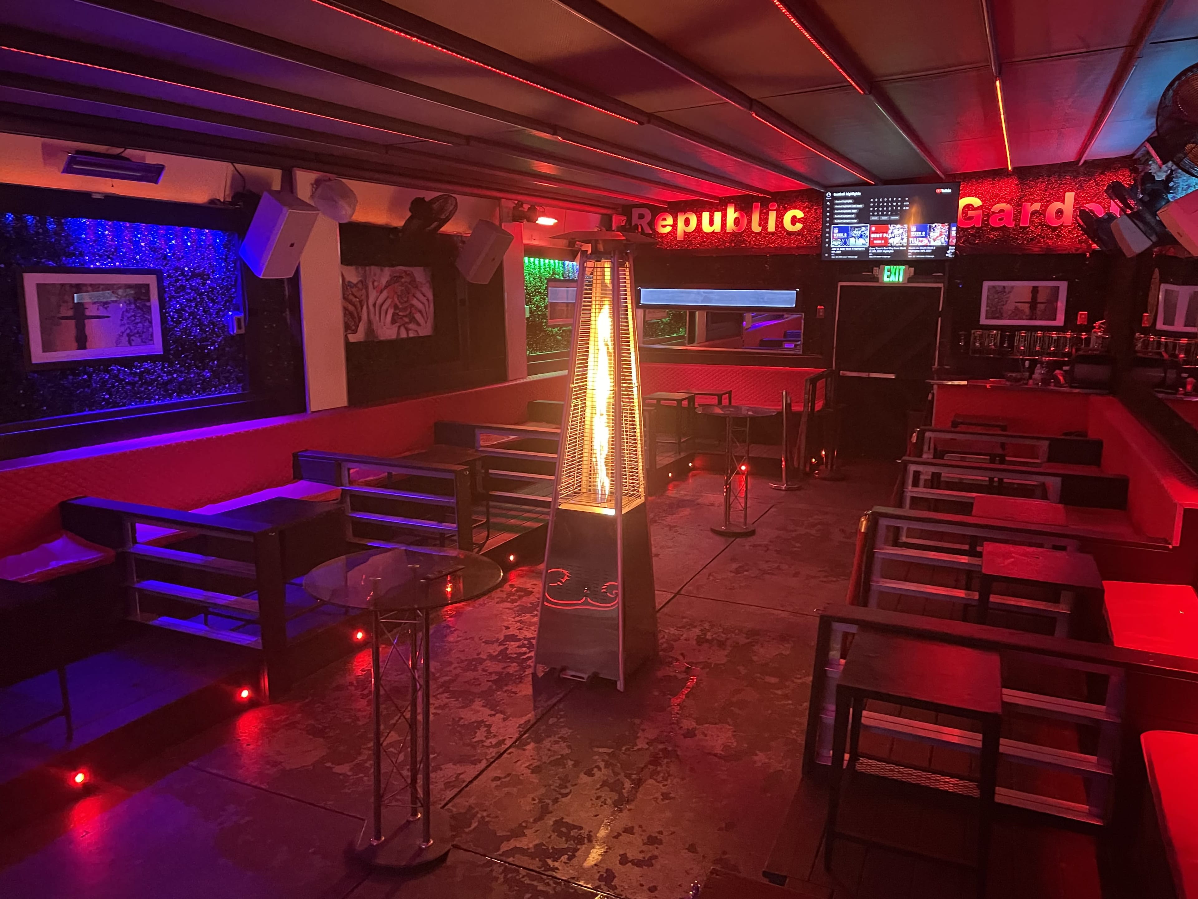 An empty lounge with red and blue lighting, featuring tables and a decorative heater in the center.

