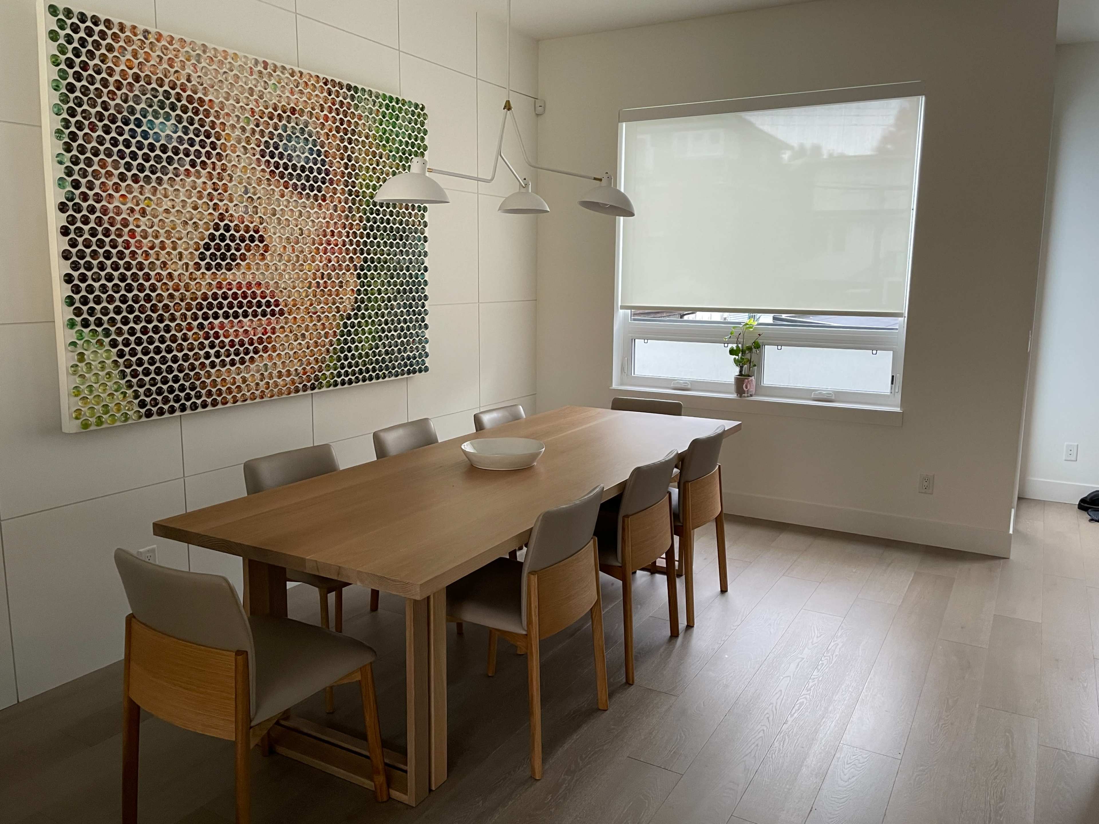 A wooden dining table with eight chairs is positioned in a well-lit room featuring a large, colorful artwork on the wall and a window with light-colored shades.