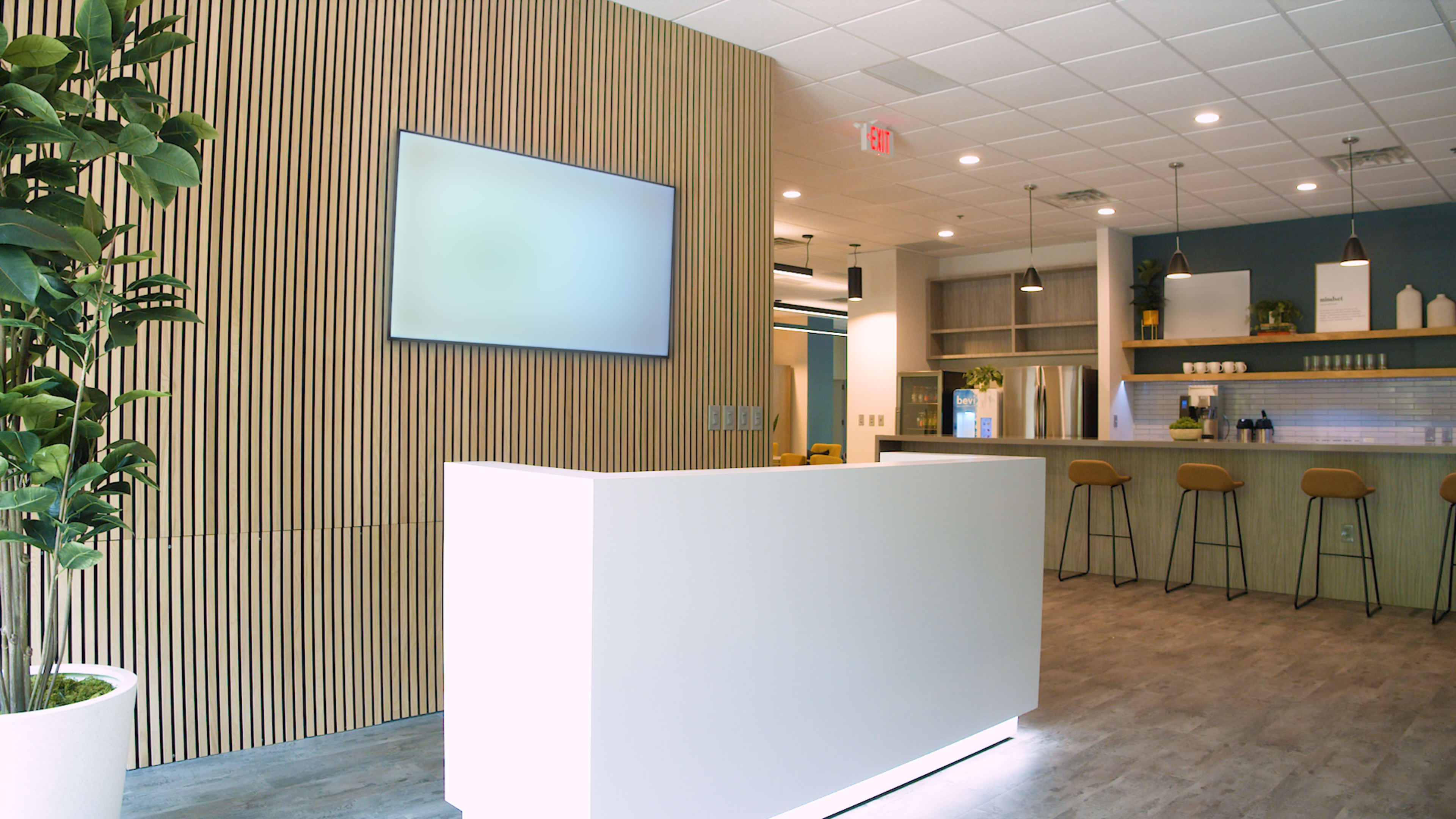 A modern reception area with a white front desk, wooden paneling on the walls, and a bar counter with stools in the background.