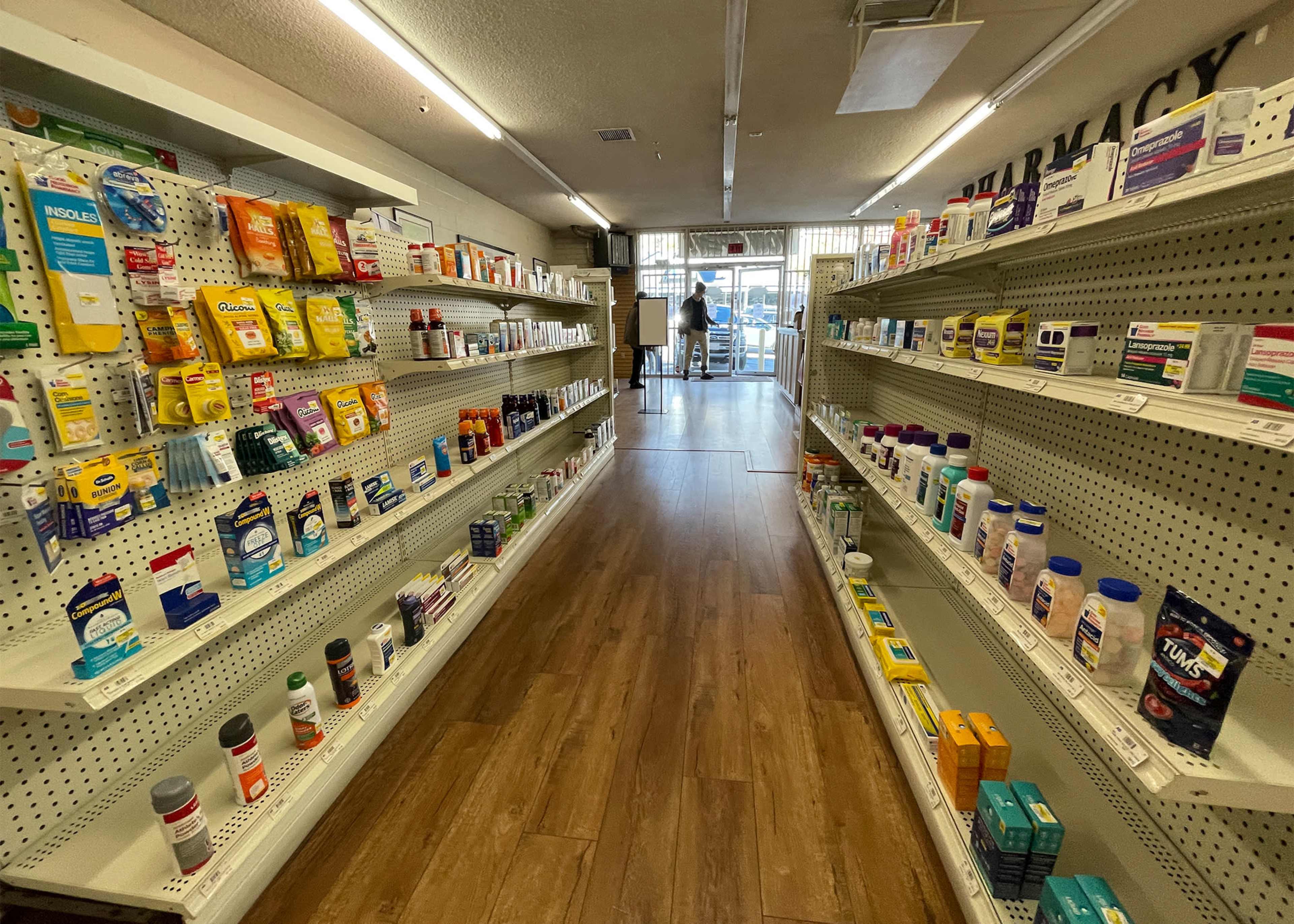 The interior of a pharmacy with shelves lined with various over-the-counter medications and health products on both sides, leading to an entrance at the far end.