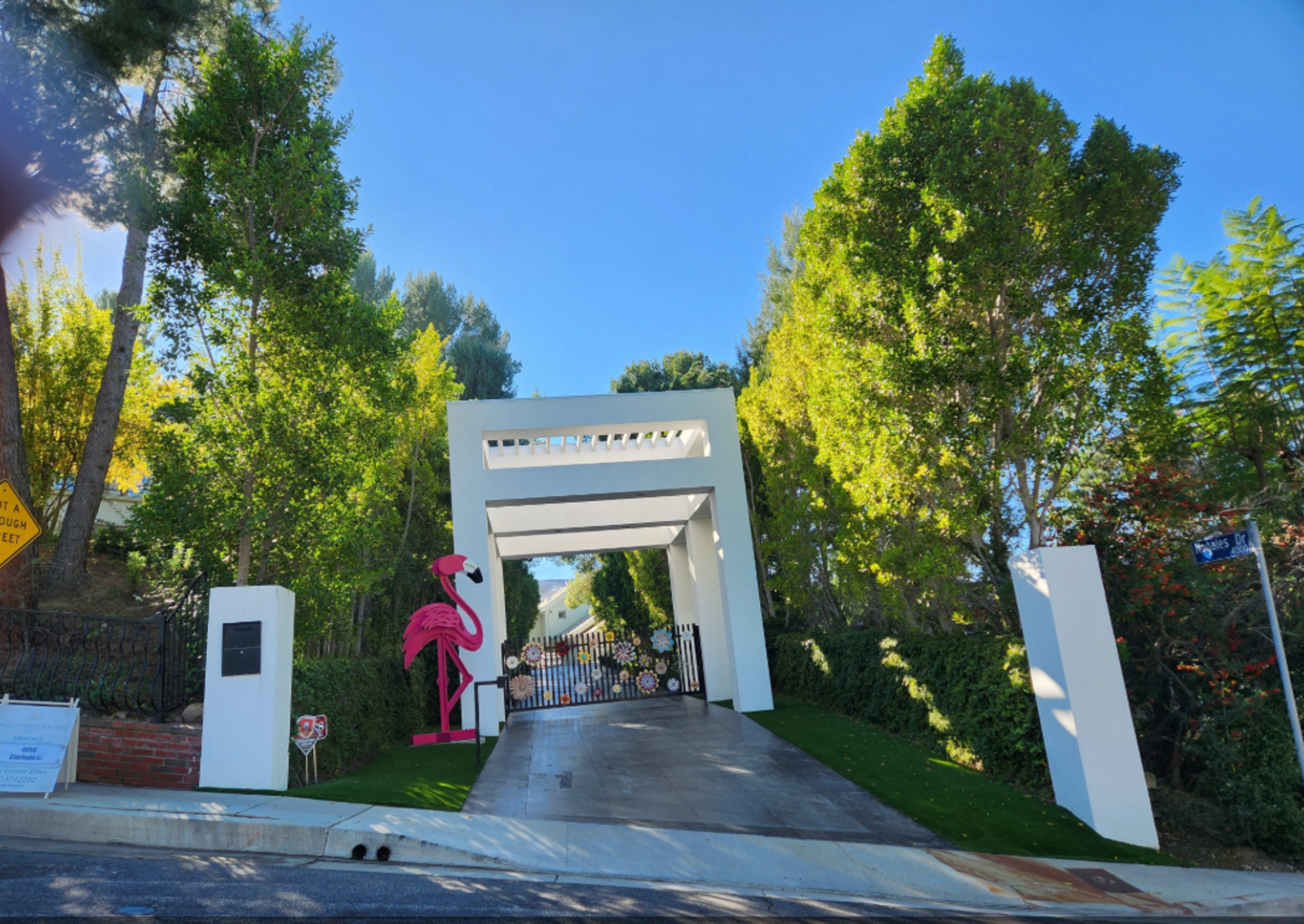 The image shows a modern gated entrance framed by tall greenery and features a pink flamingo decoration at the side.