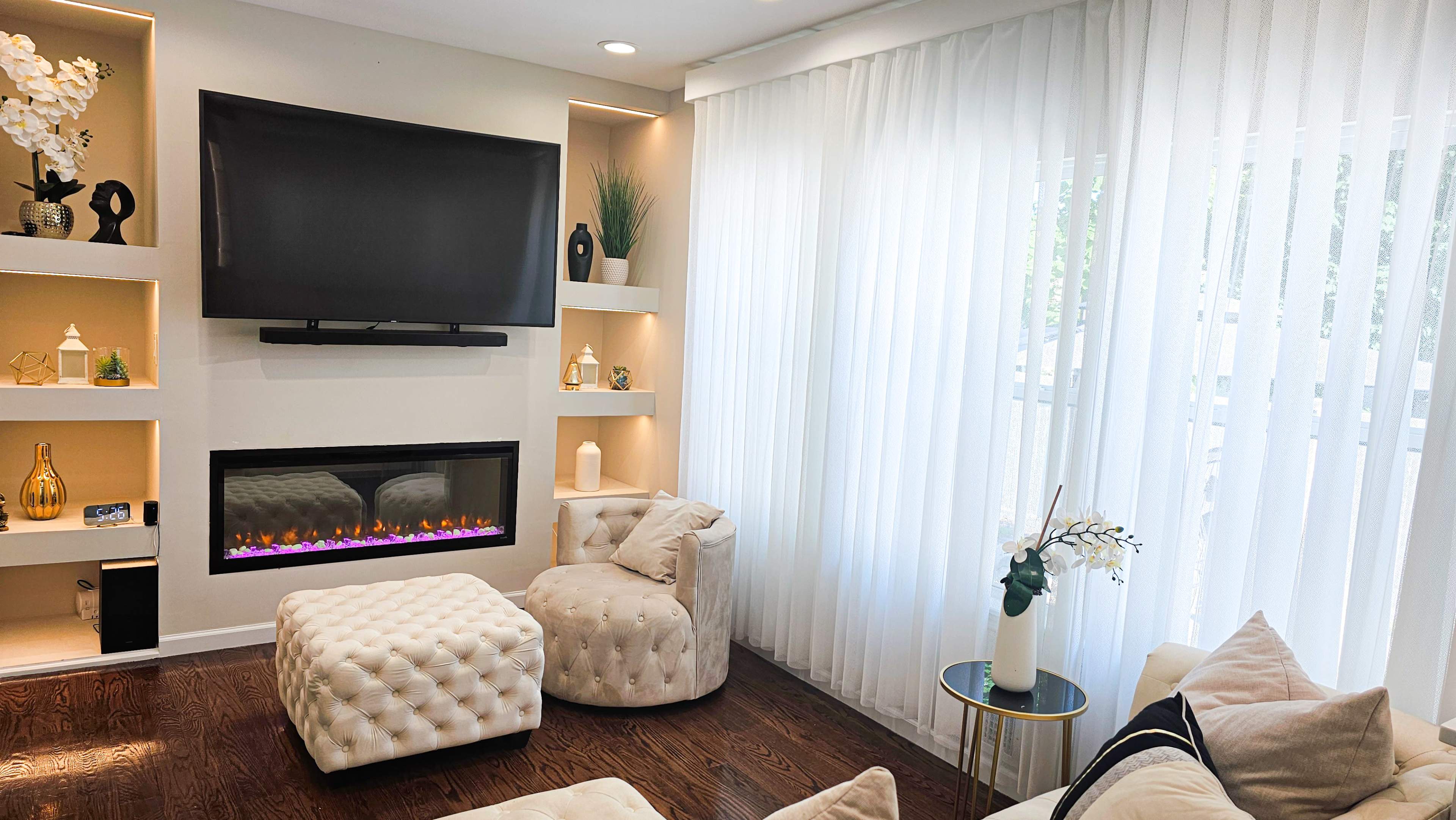 A modern living room with a mounted television, a wall-mounted electric fireplace, and light-colored furniture against a backdrop of sheer curtains.