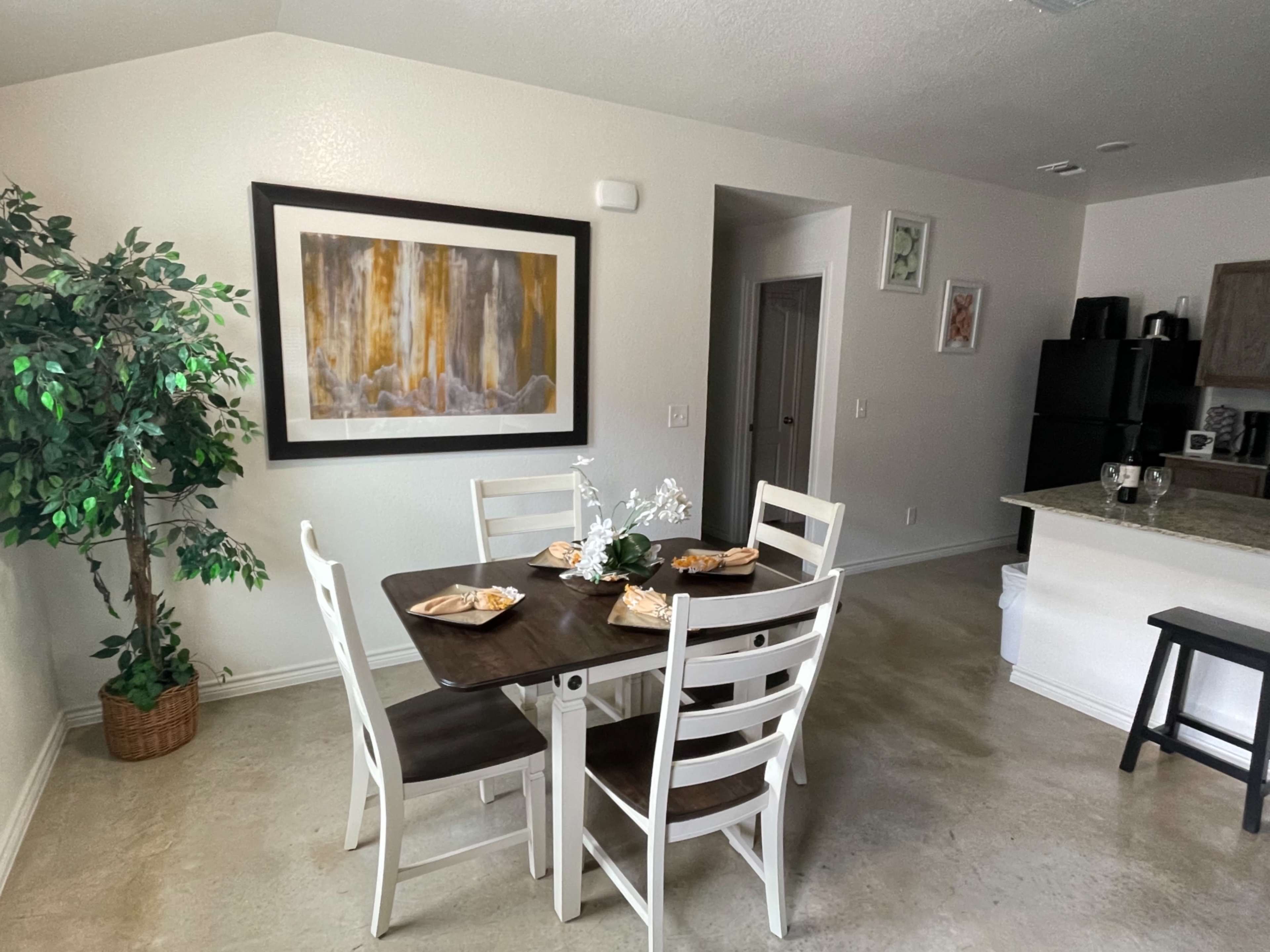 A dining area features a table set for four, surrounded by chairs, with a large framed artwork and a small artificial tree in the corner.