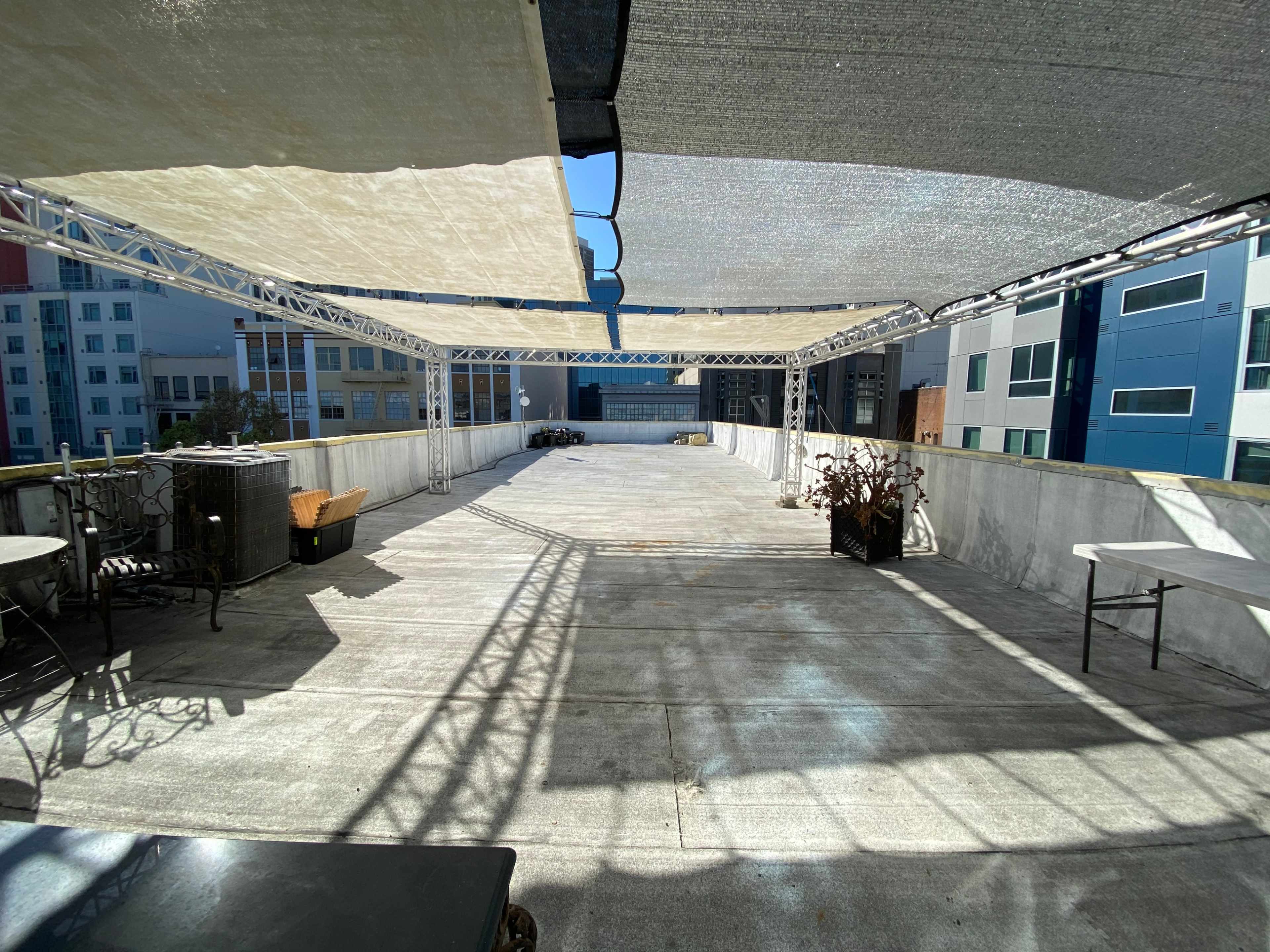 An empty rooftop space with overhead fabric shades and concrete flooring, surrounded by buildings.