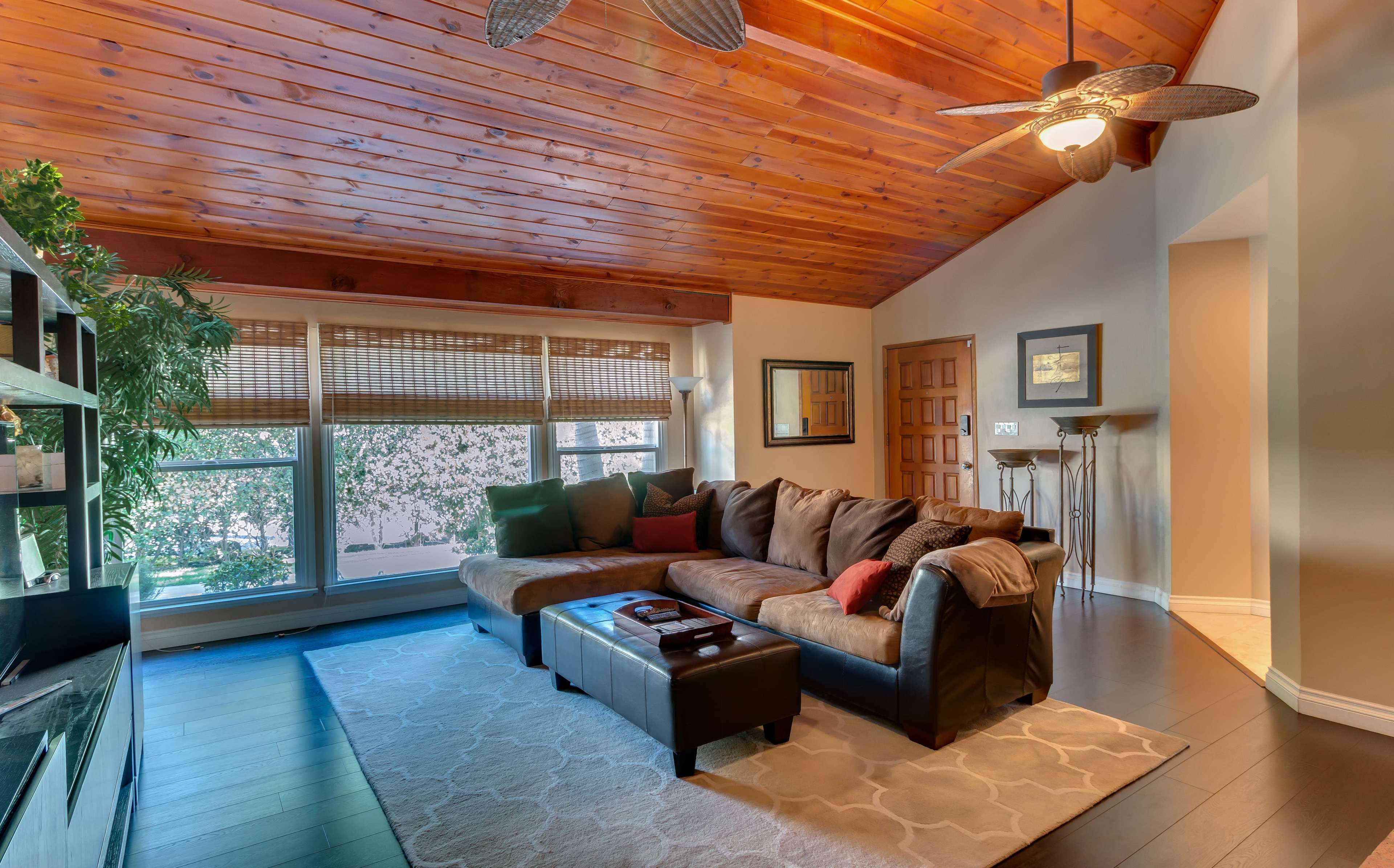 The living room features a large brown sectional sofa, a coffee table, and wooden ceiling beams with a ceiling fan, illuminated by natural light from the large windows.