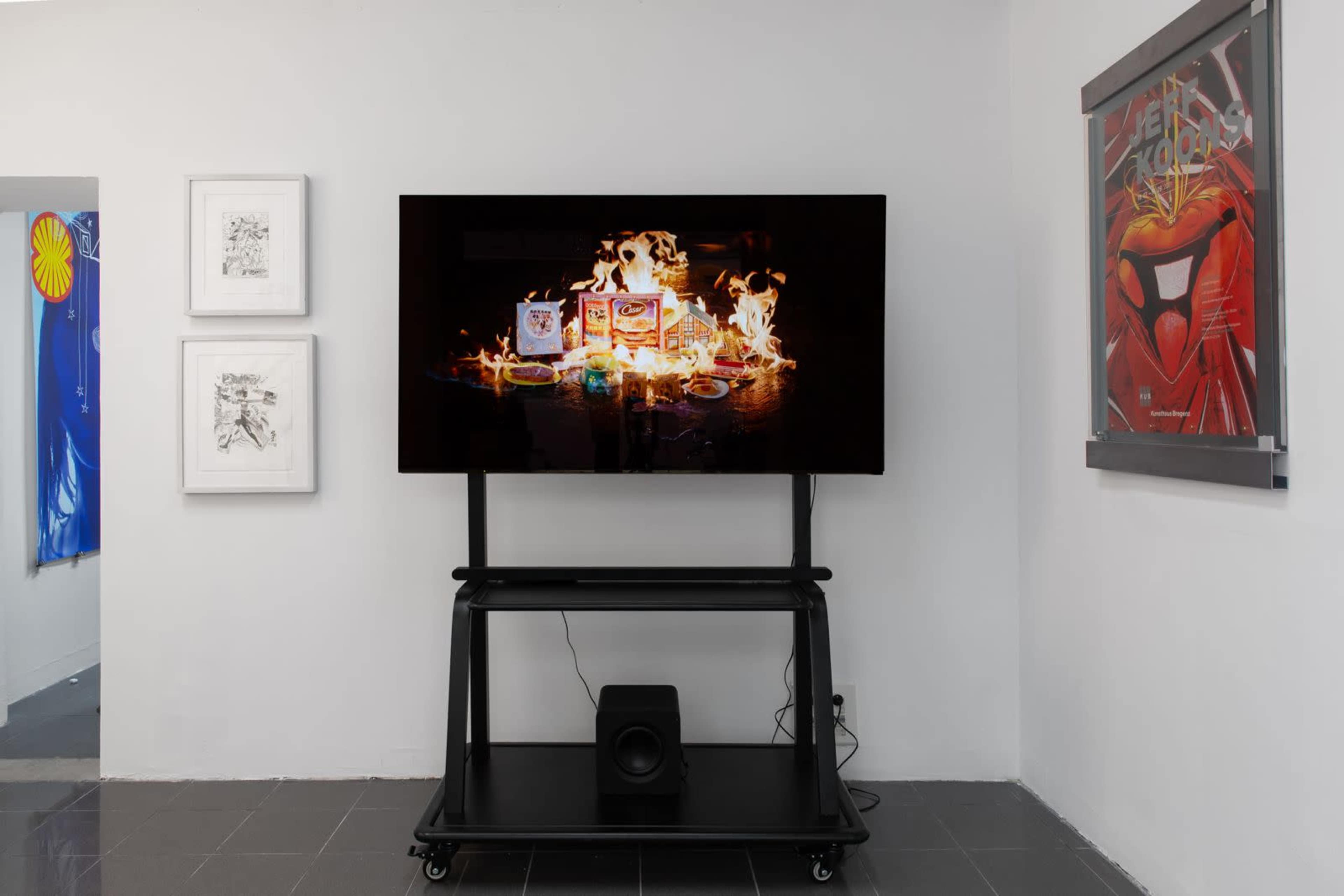 The image shows a large television displaying an artwork featuring various colorful products surrounded by flames, positioned on a stand in a white-walled room with framed artworks on either side.