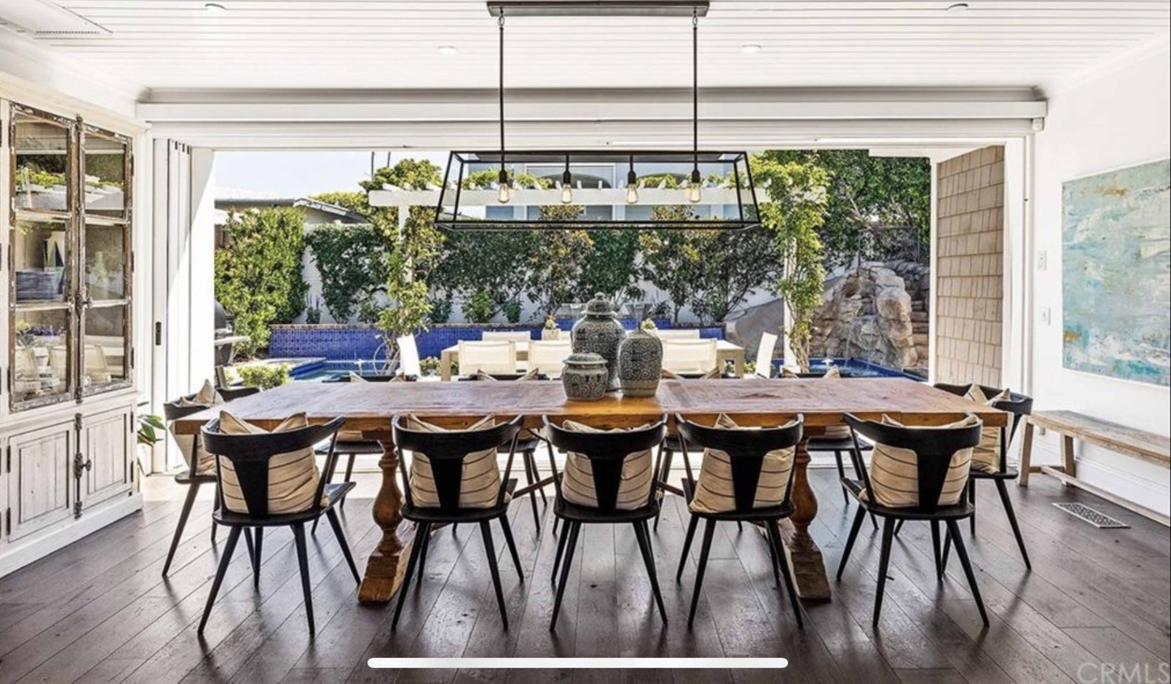 A large wooden dining table with black chairs is set within an open interior space that overlooks a pool area and garden through glass sliding doors.