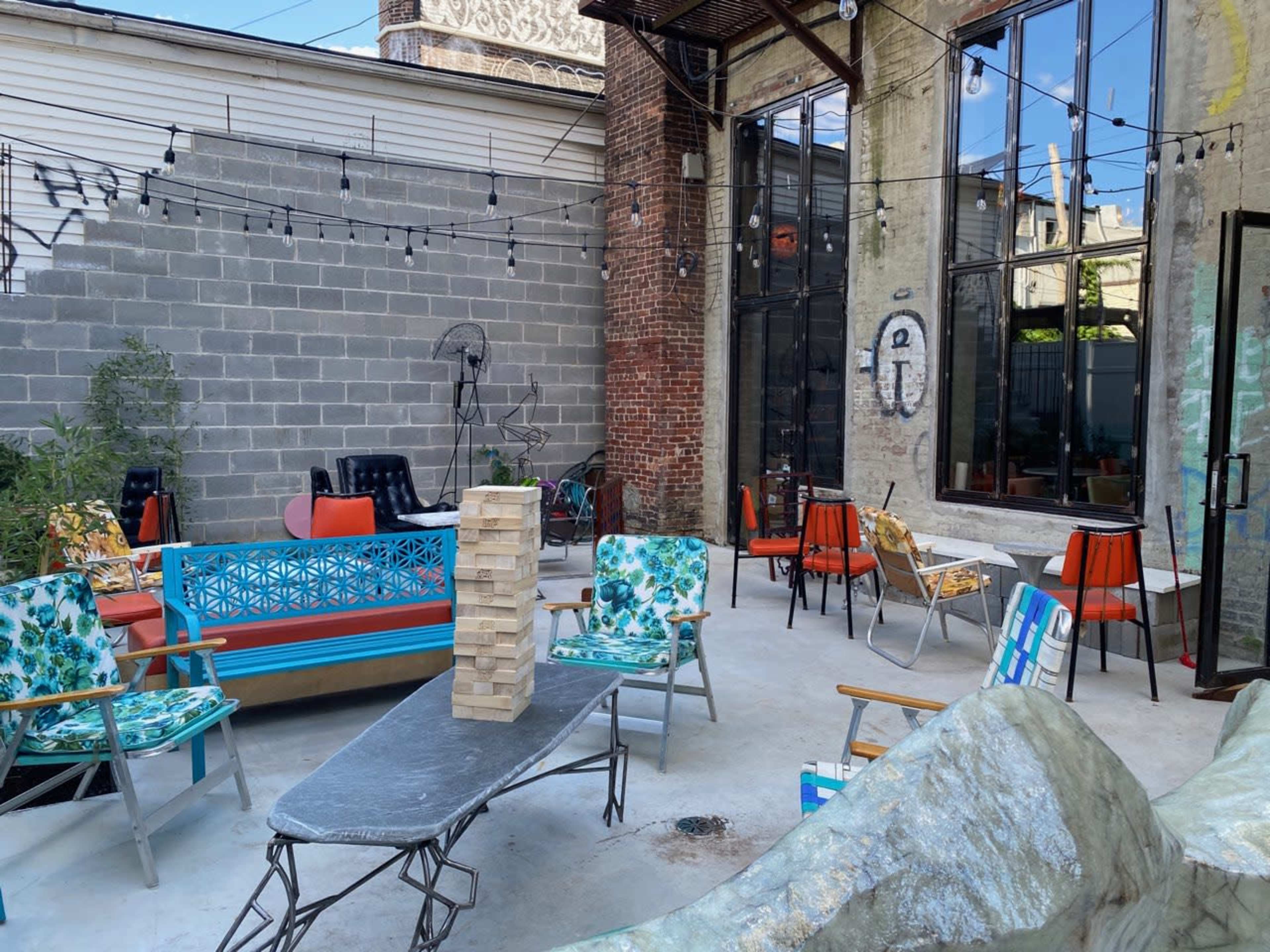 The image depicts a vibrant outdoor seating area with colorful chairs, a game of Jenga, and string lights strung across a rustic, industrial setting.