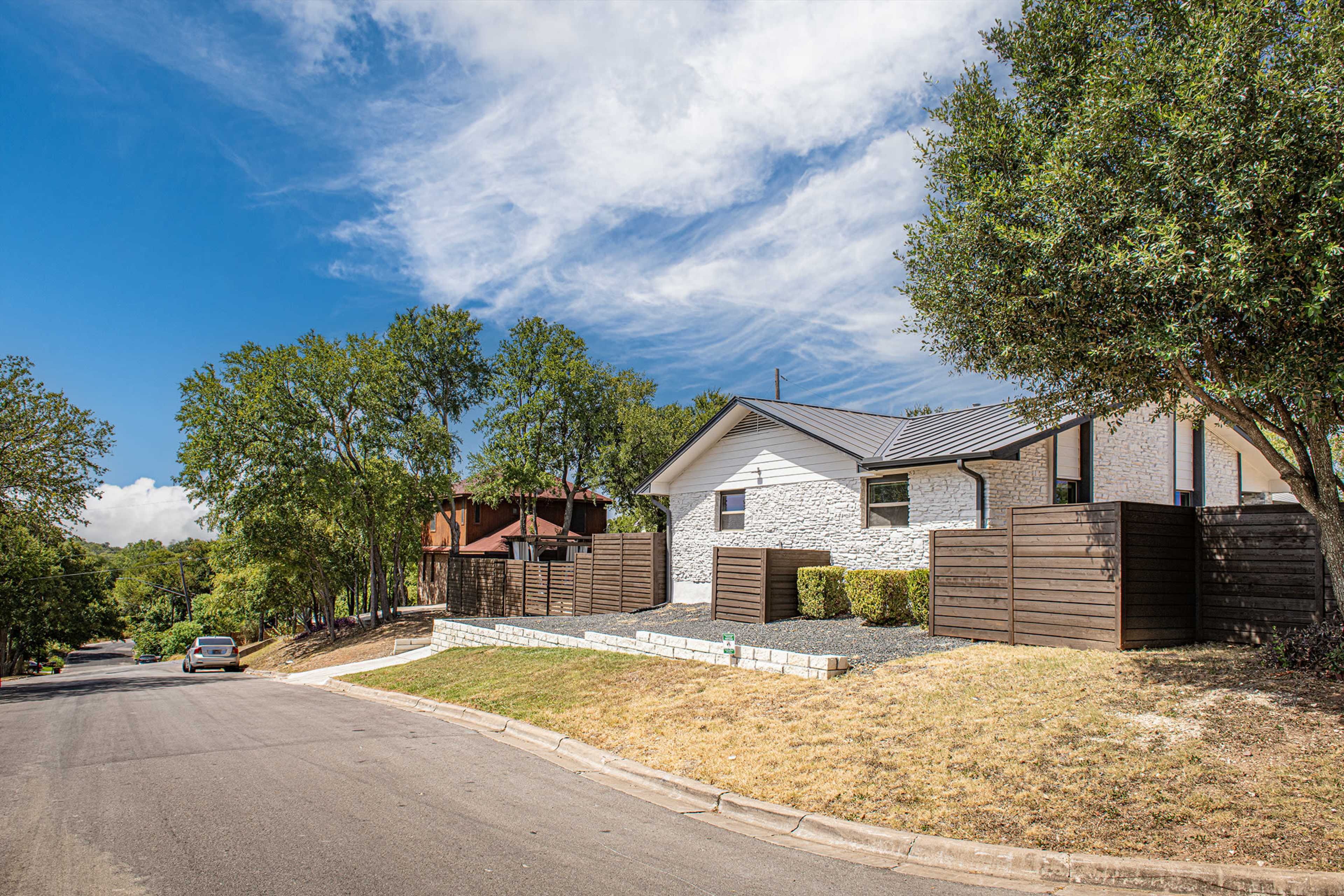 A single-story house with a stone facade and wooden fencing sits on a sloped street lined with trees against a partly cloudy sky.