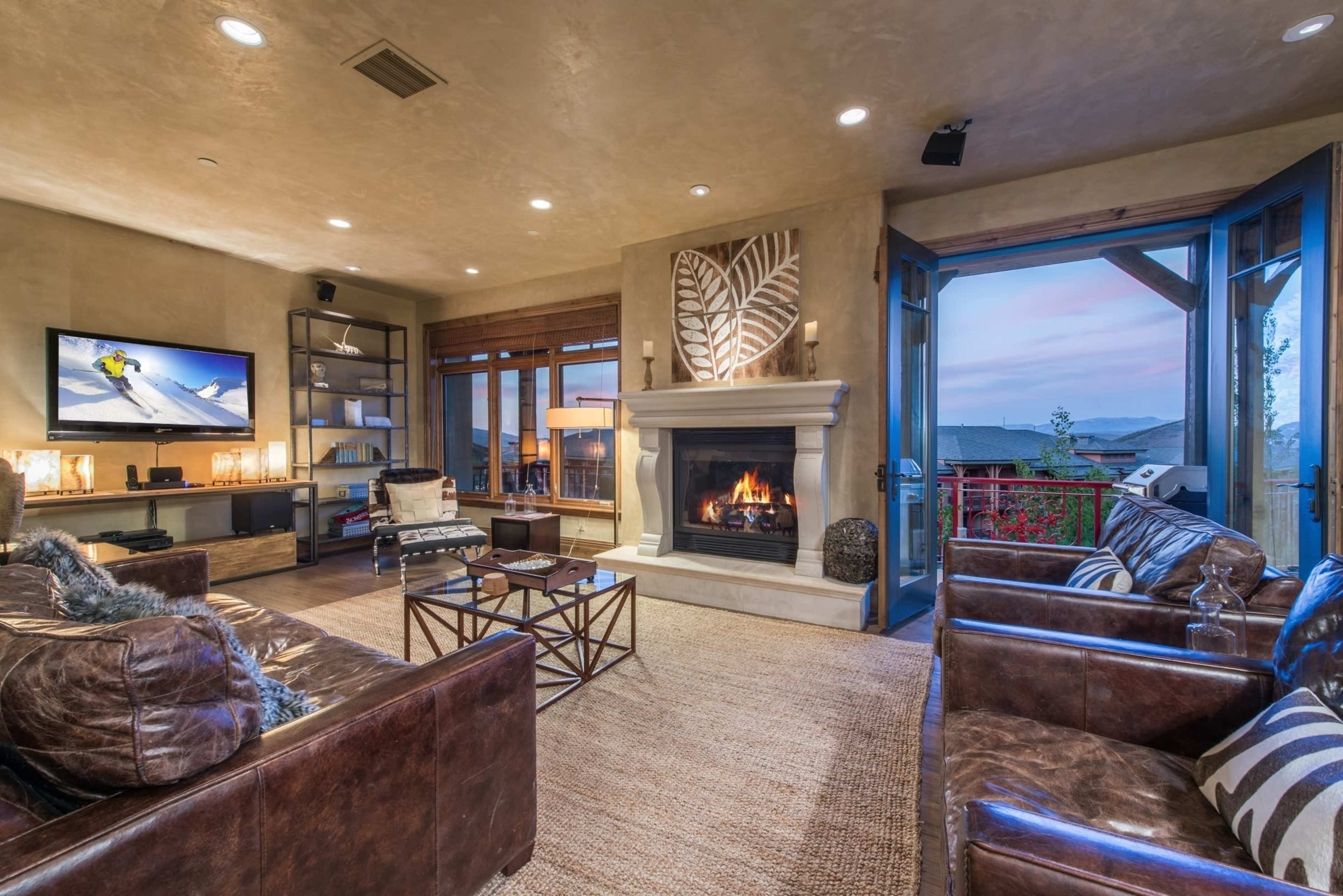 A modern living room with a fireplace, leather seating, a television, and large windows that open to a scenic view.