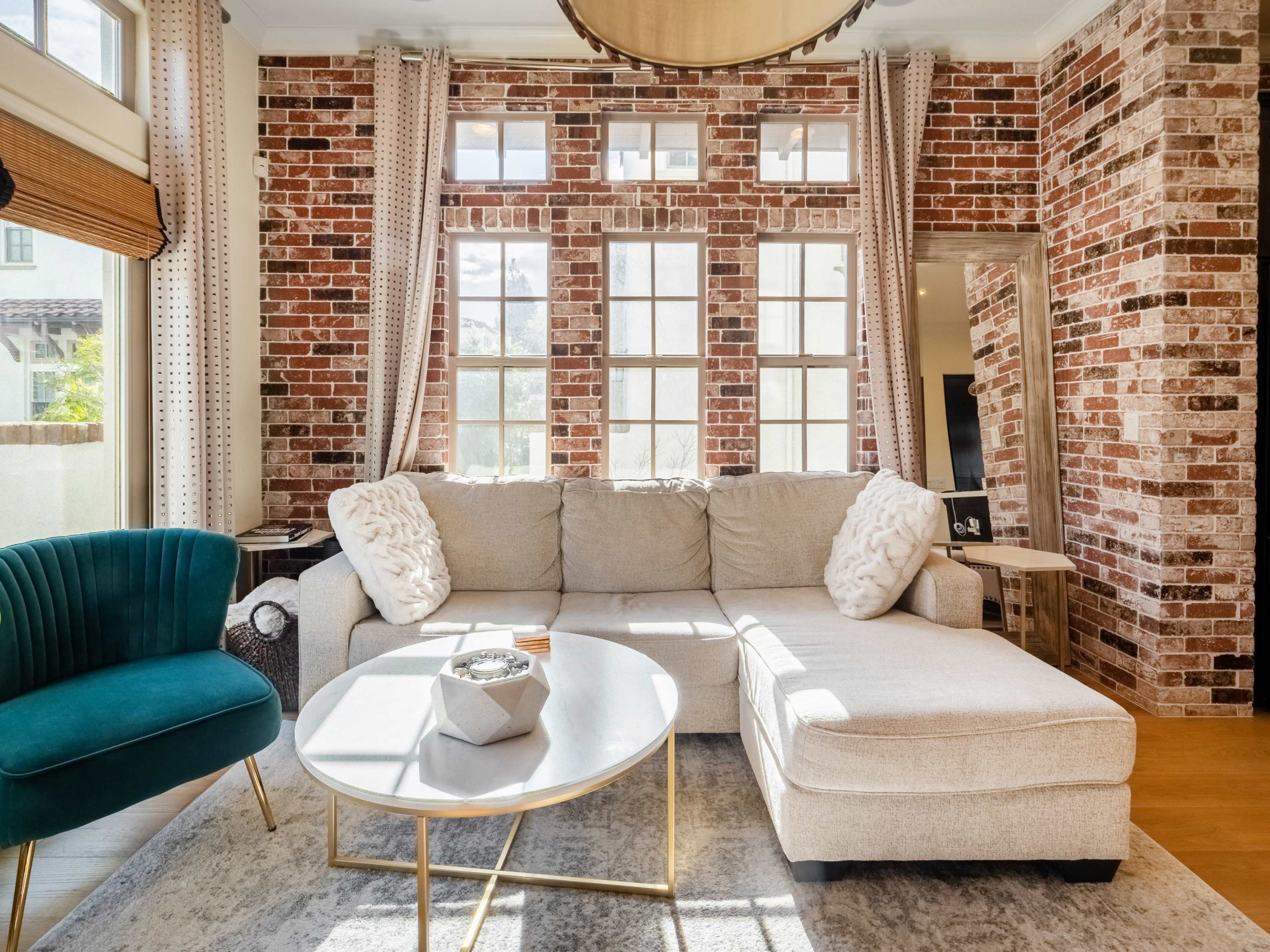 The image shows a modern living room with a sectional sofa, a round coffee table, and large windows framed by brick walls.