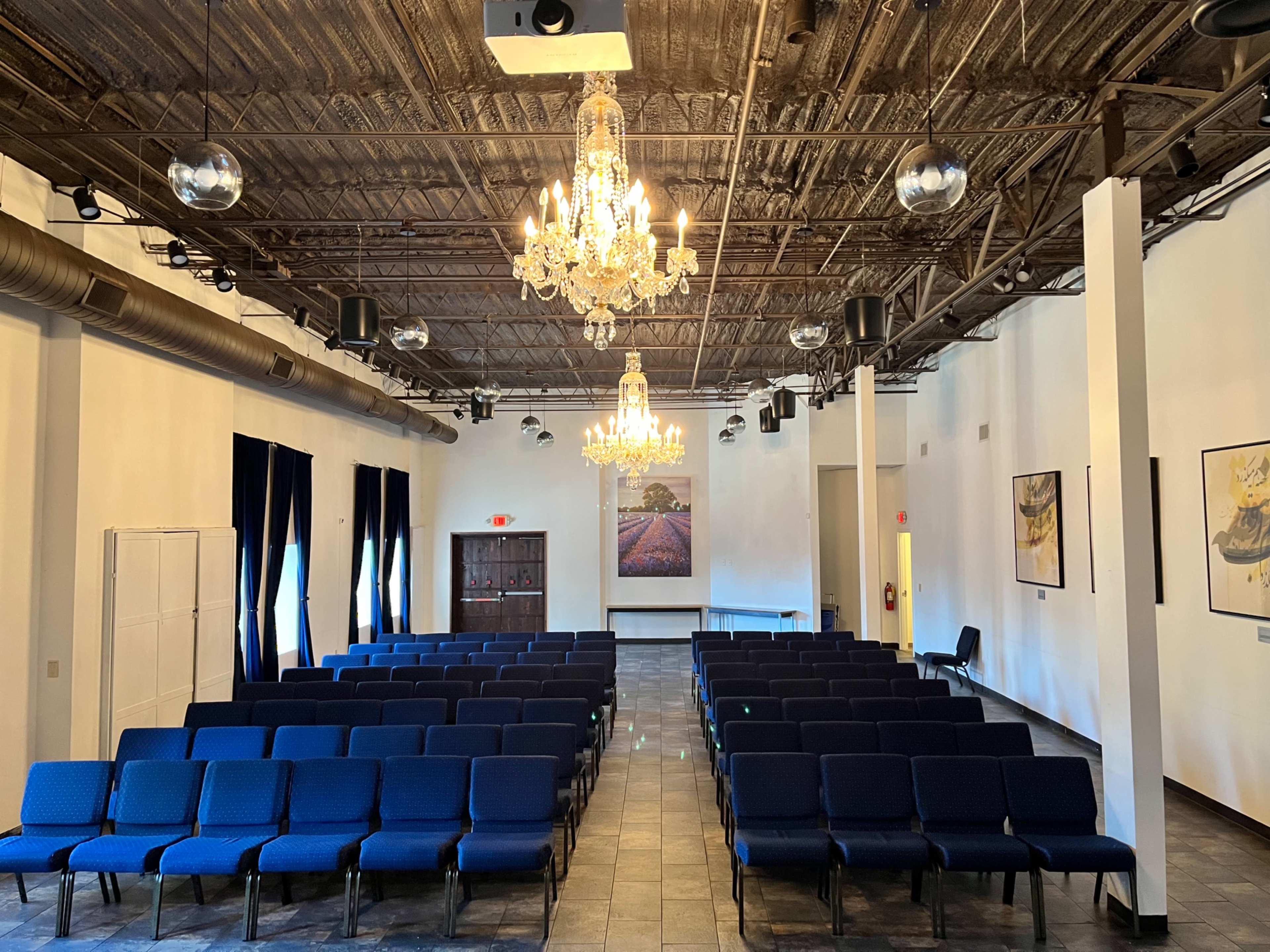 The image shows an empty room with rows of blue chairs, a chandelier, and artwork on the walls.
