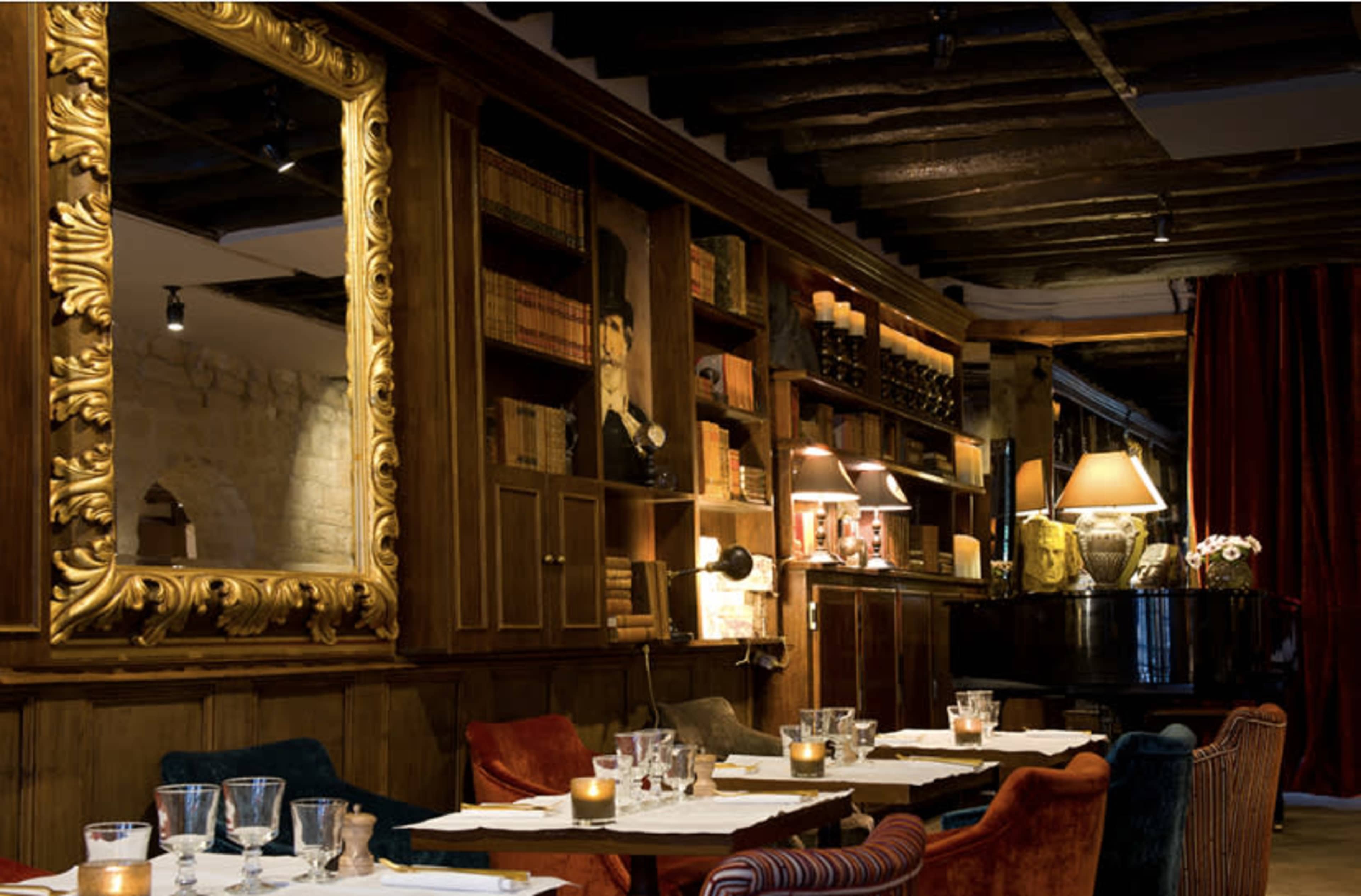 An elegantly decorated restaurant interior featuring wooden shelving filled with books, a grand piano, and a large ornate mirror reflecting the space.