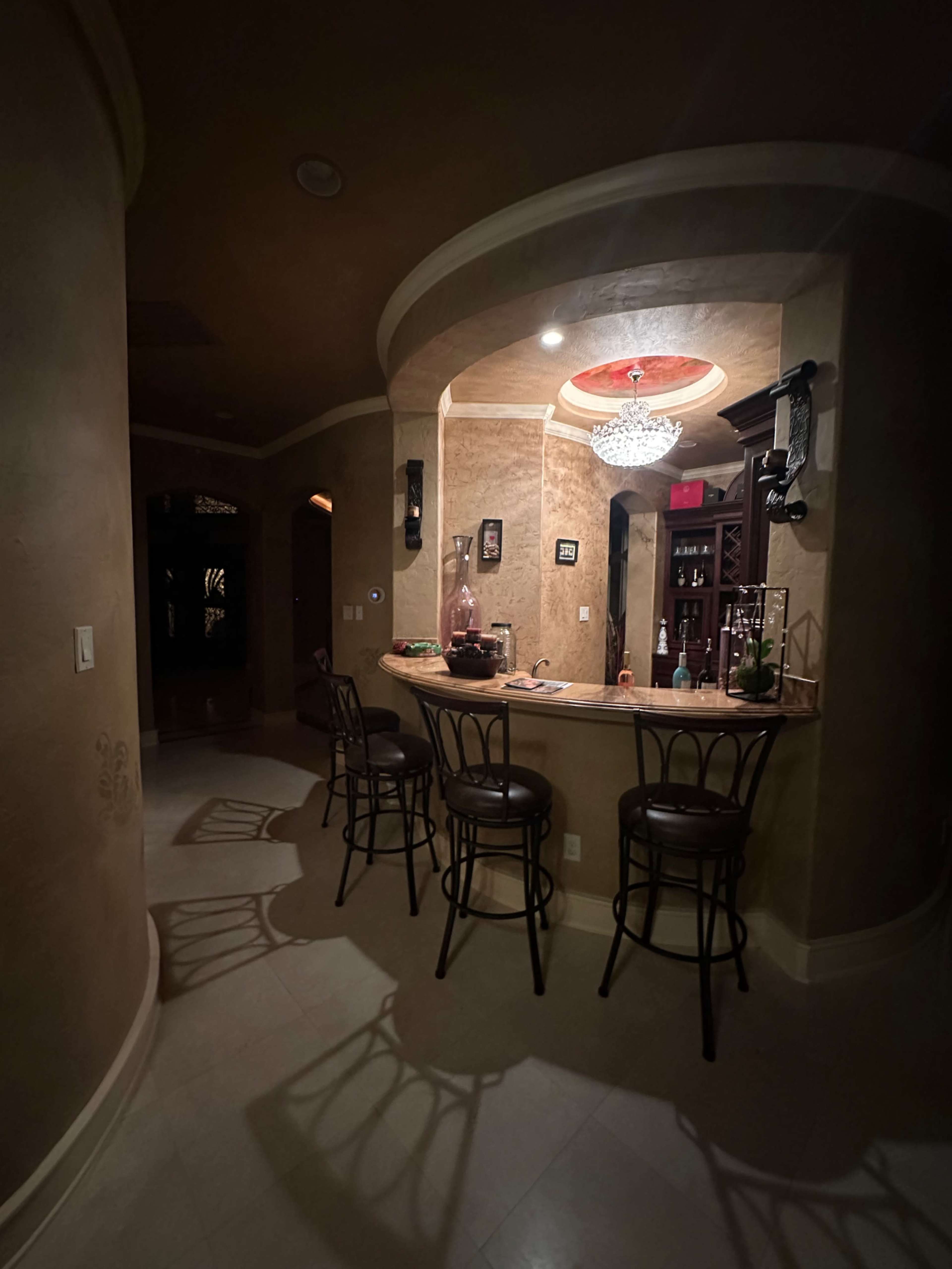 The image shows a dimly lit bar area with four stools and a curved counter, adorned with a chandelier and decorated walls.