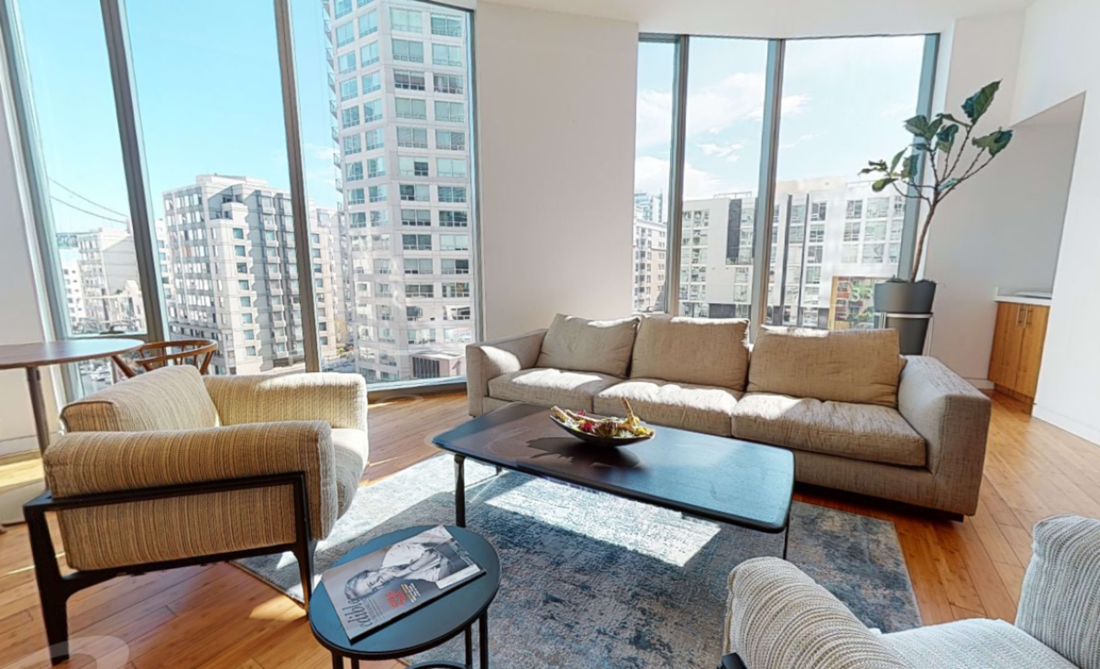 A bright living room features large windows with city skyline views, modern furniture, and a small dining area.