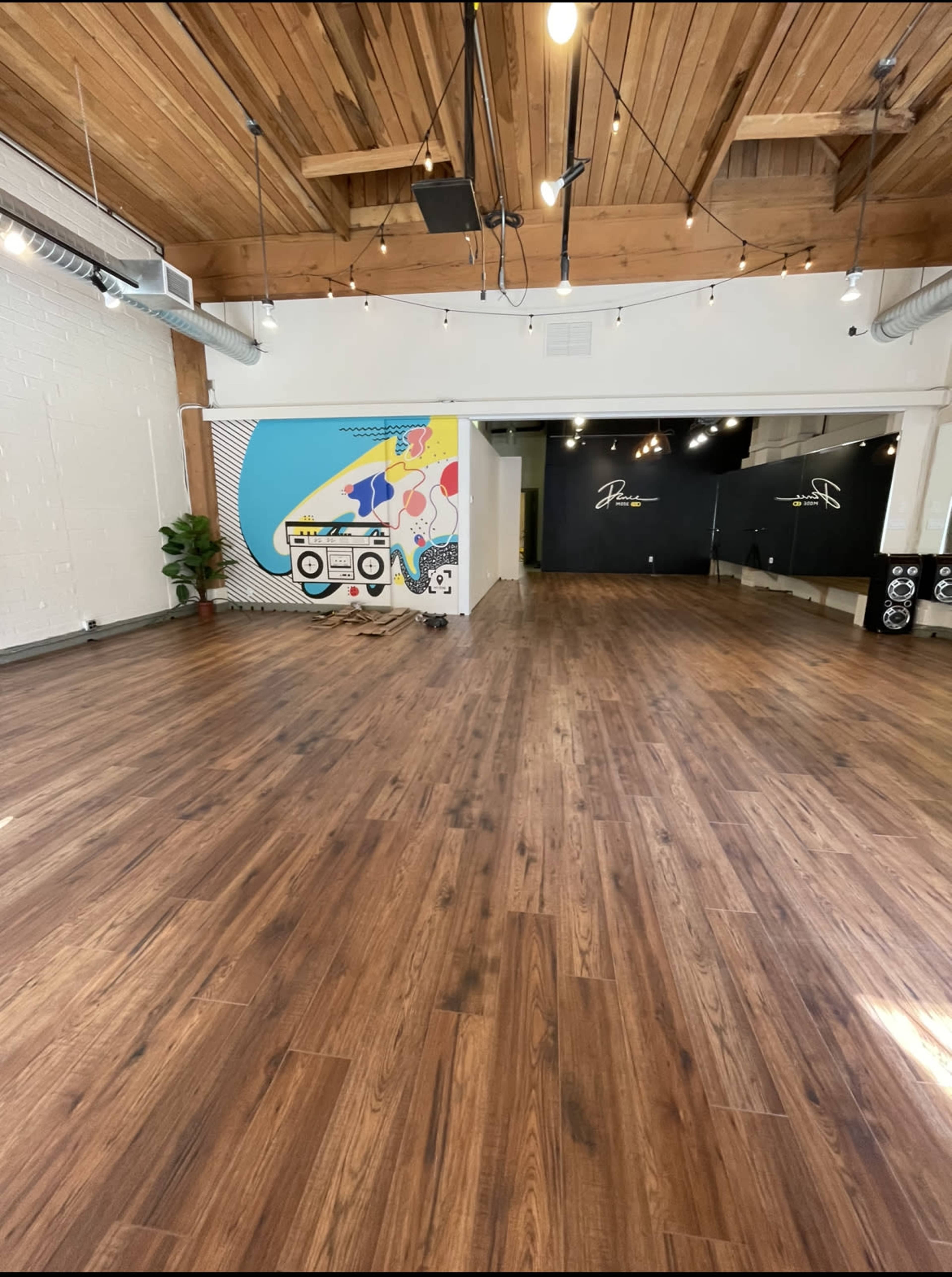 A spacious, wooden-floored studio featuring a colorful mural on one wall and a dark wall with lighting at the far end.