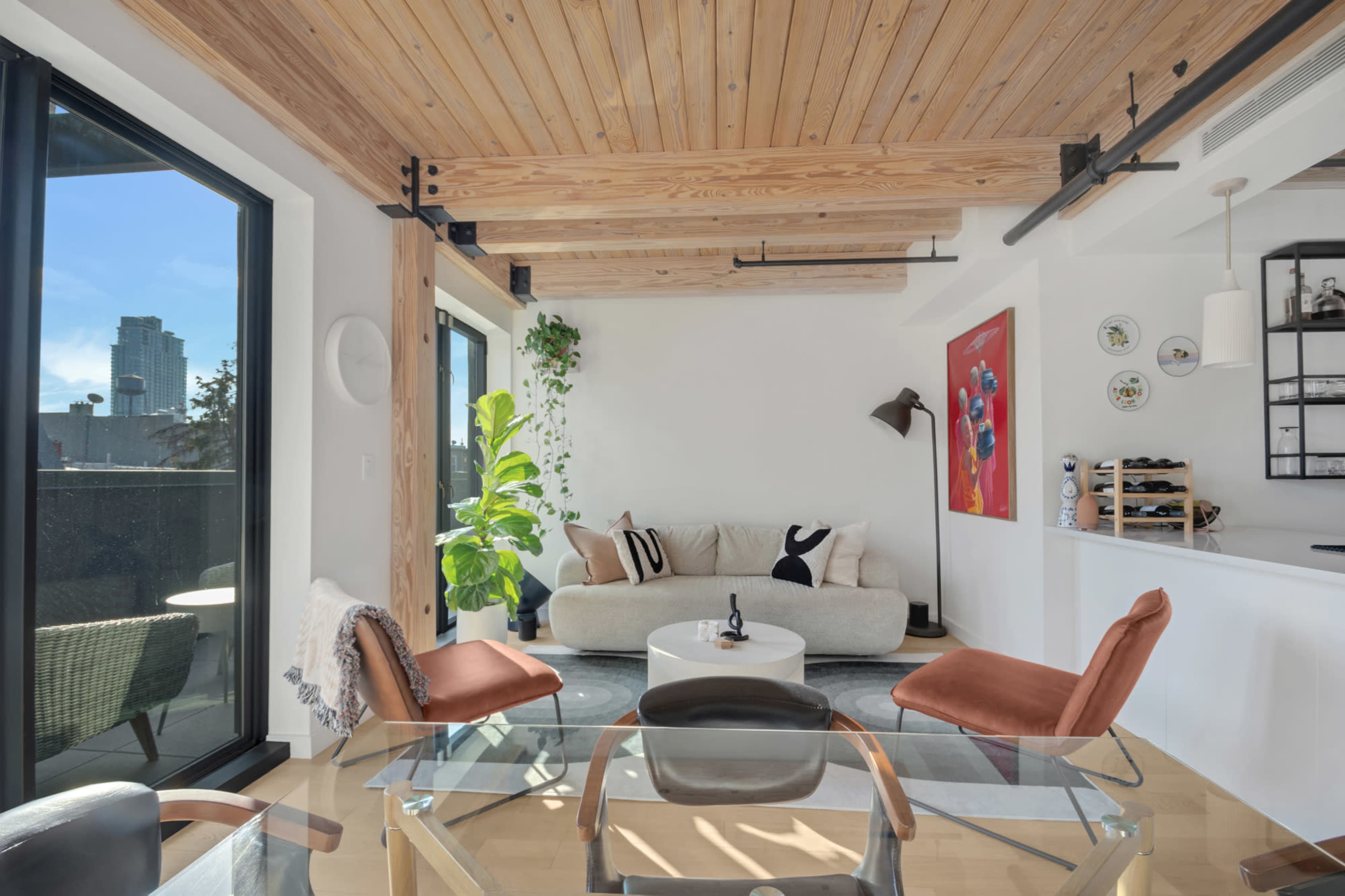 The image shows a modern living room featuring a light-colored sofa, a glass dining table with black chairs, and large windows with a view of a city skyline.