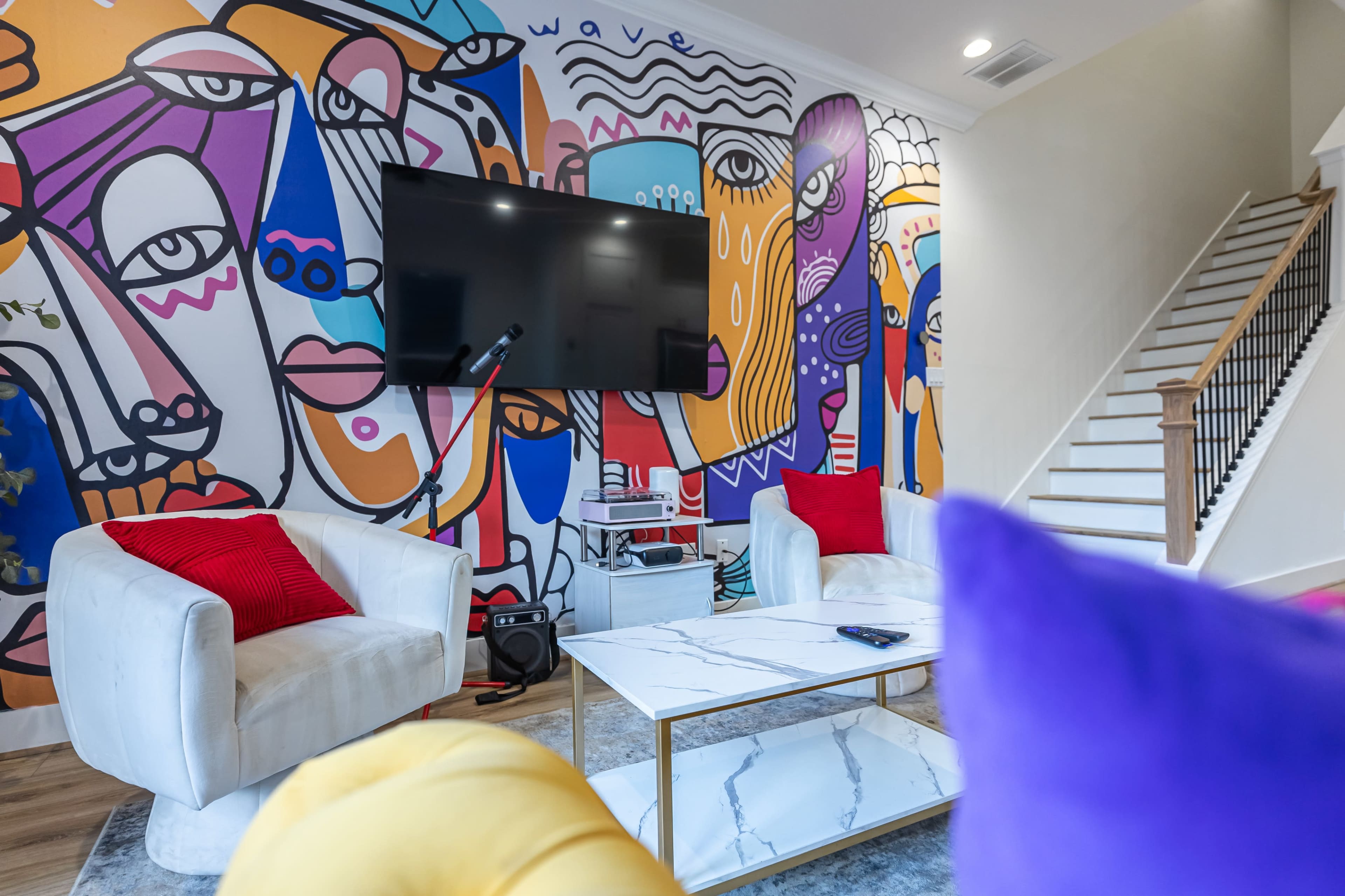A colorful mural featuring abstract faces adorns the wall of a modern living room that includes two white chairs, a glass coffee table, and a large flat-screen TV.