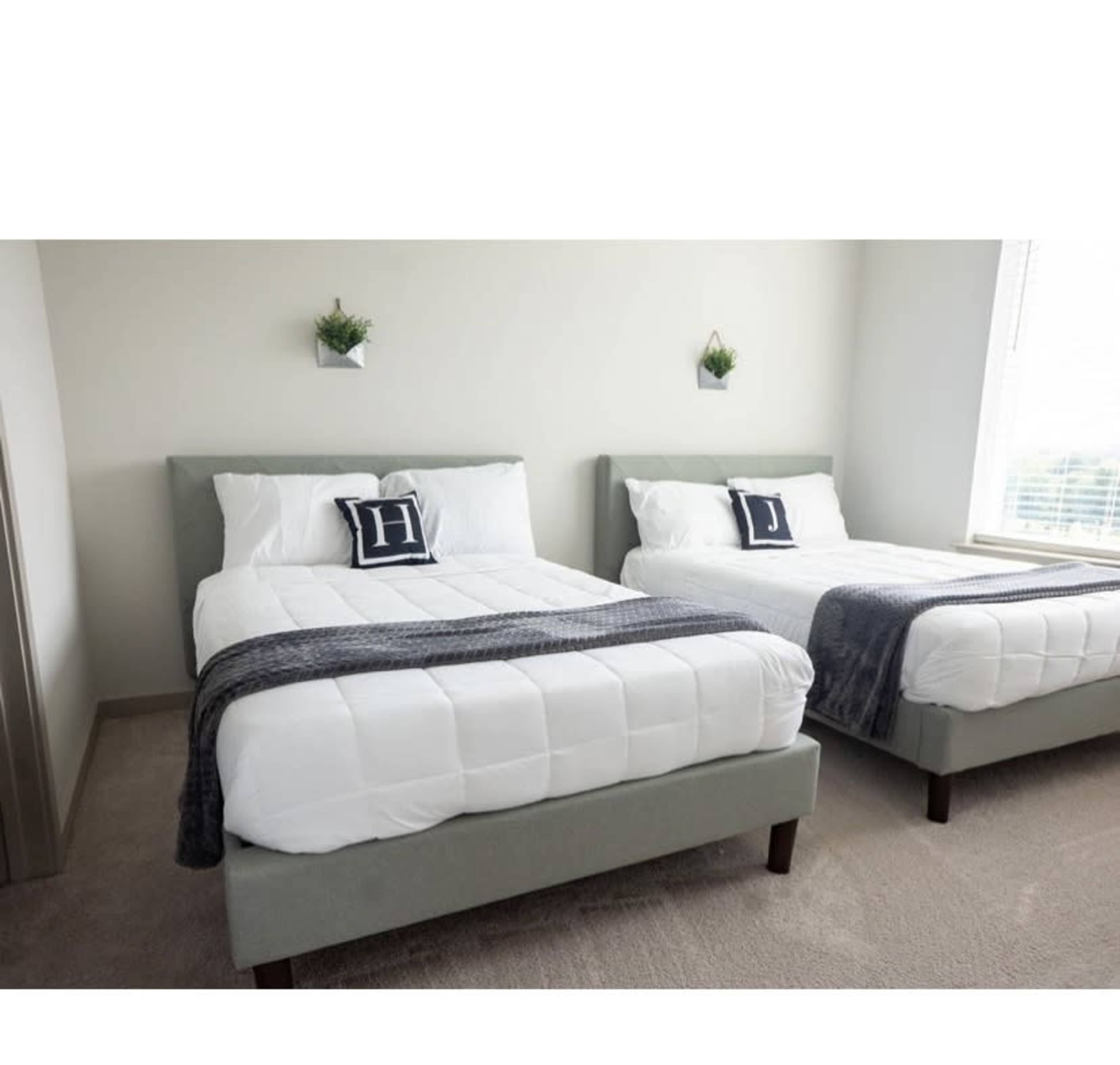 A room with two neatly made beds, each with white bedding and decorative pillows featuring the letters "H" and "J."