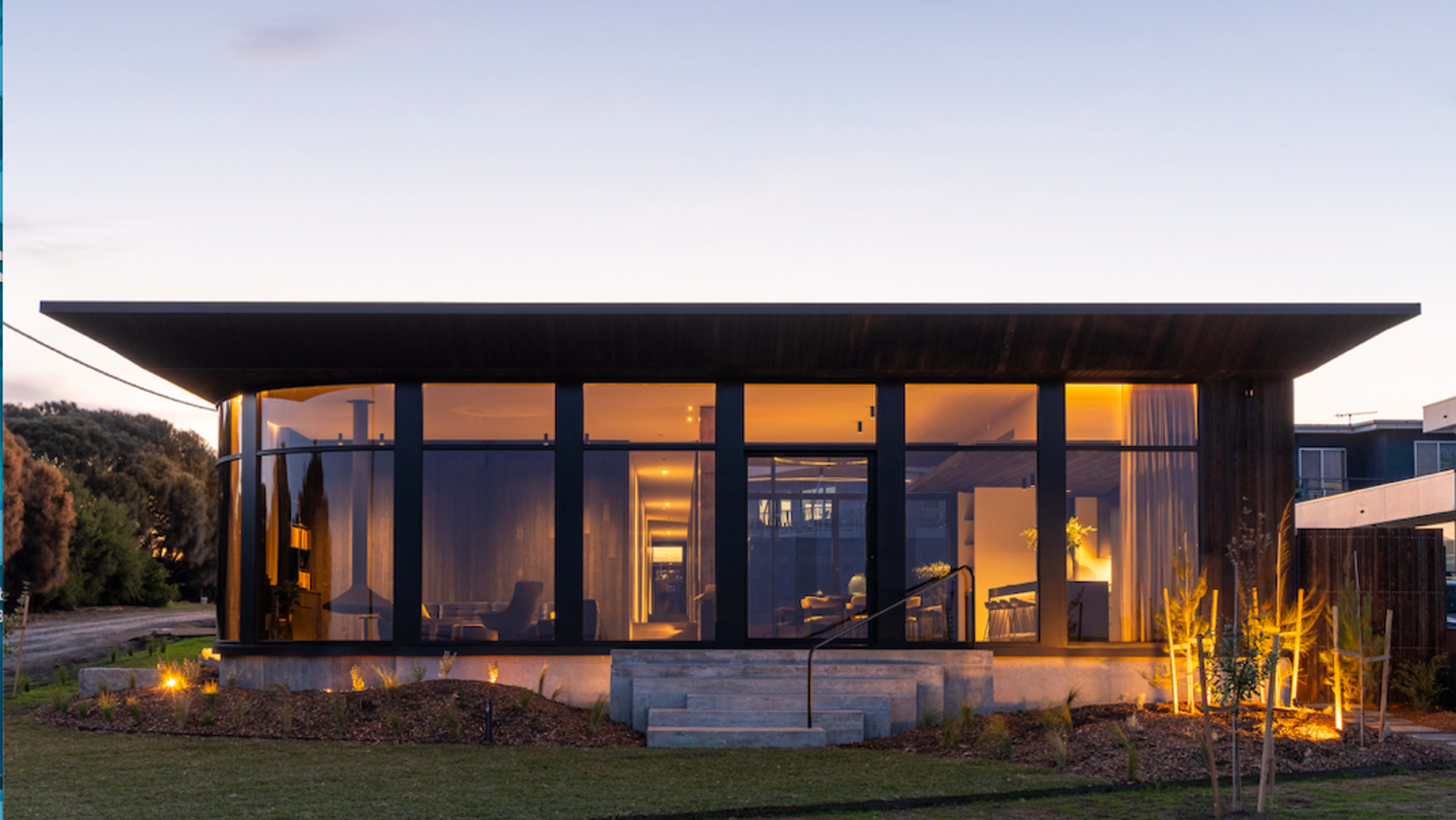 A modern house with extensive glass windows and a smooth roofline sits in a landscaped yard, illuminated warmly at dusk.