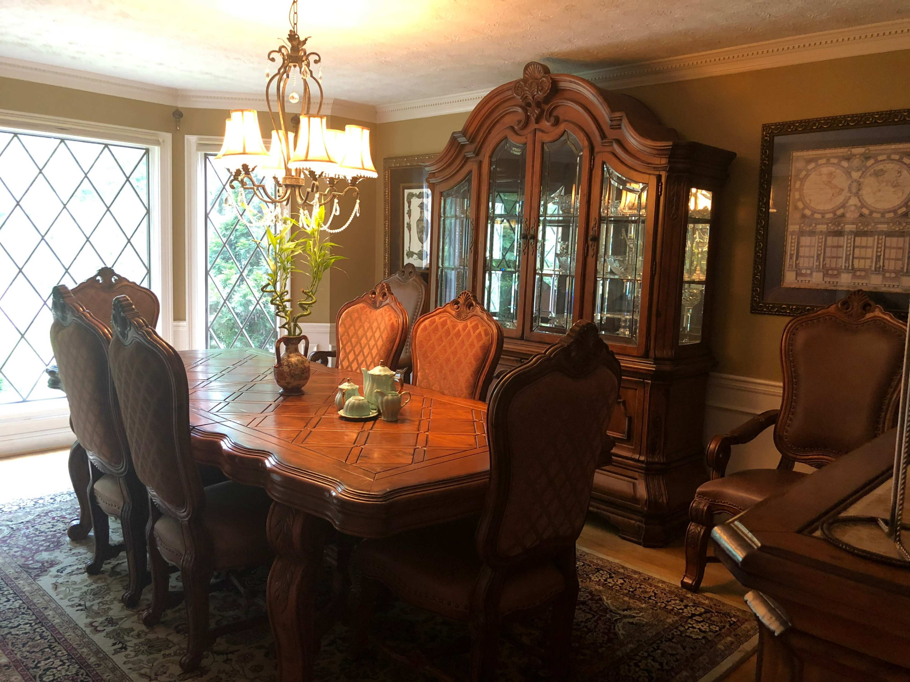 The image shows a dining room featuring a large wooden table with six upholstered chairs, a china cabinet, and a chandelier.