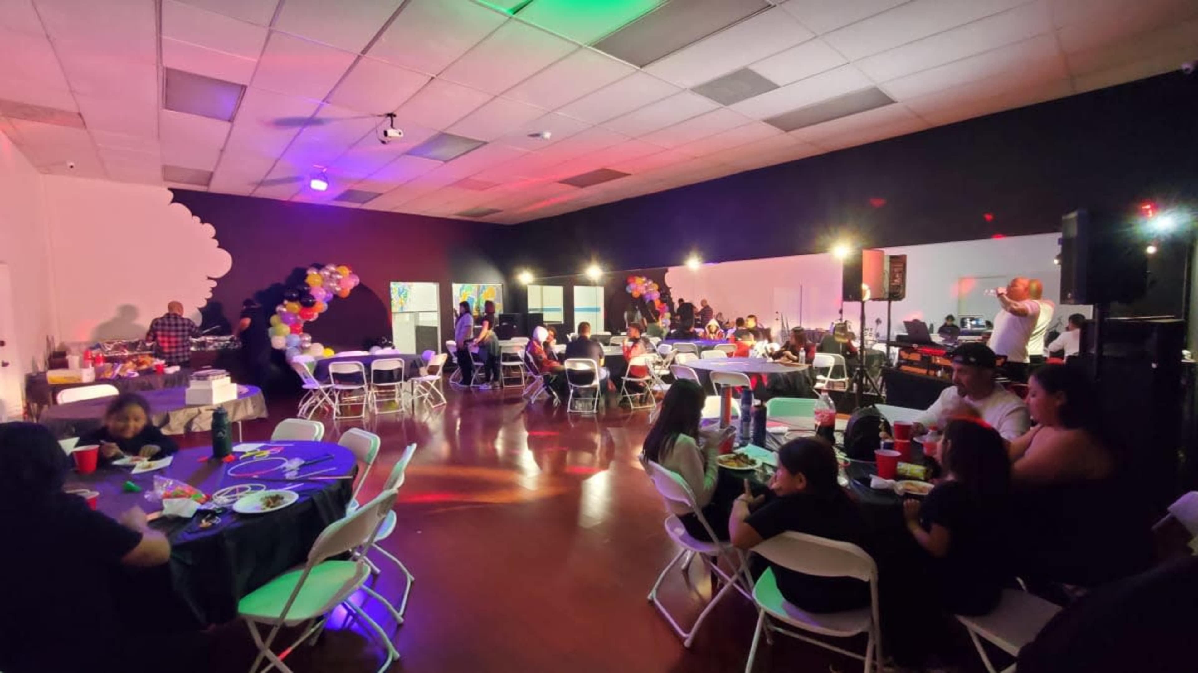 A large event space is filled with people sitting at tables, with colorful decorations and lighting enhancing the atmosphere.