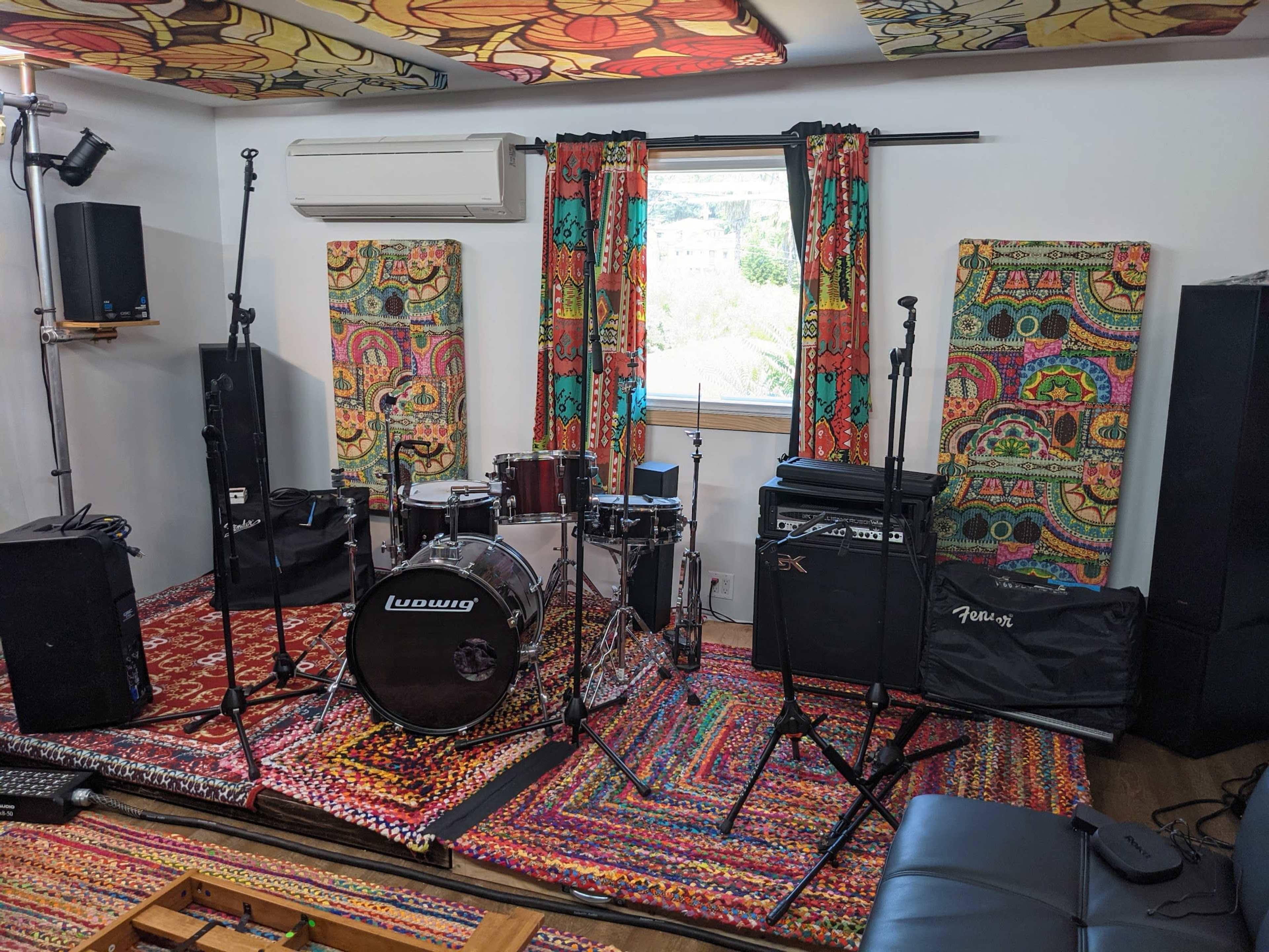 A music rehearsal space with a drum set, amplifiers, microphones, and colorful patterned curtains.