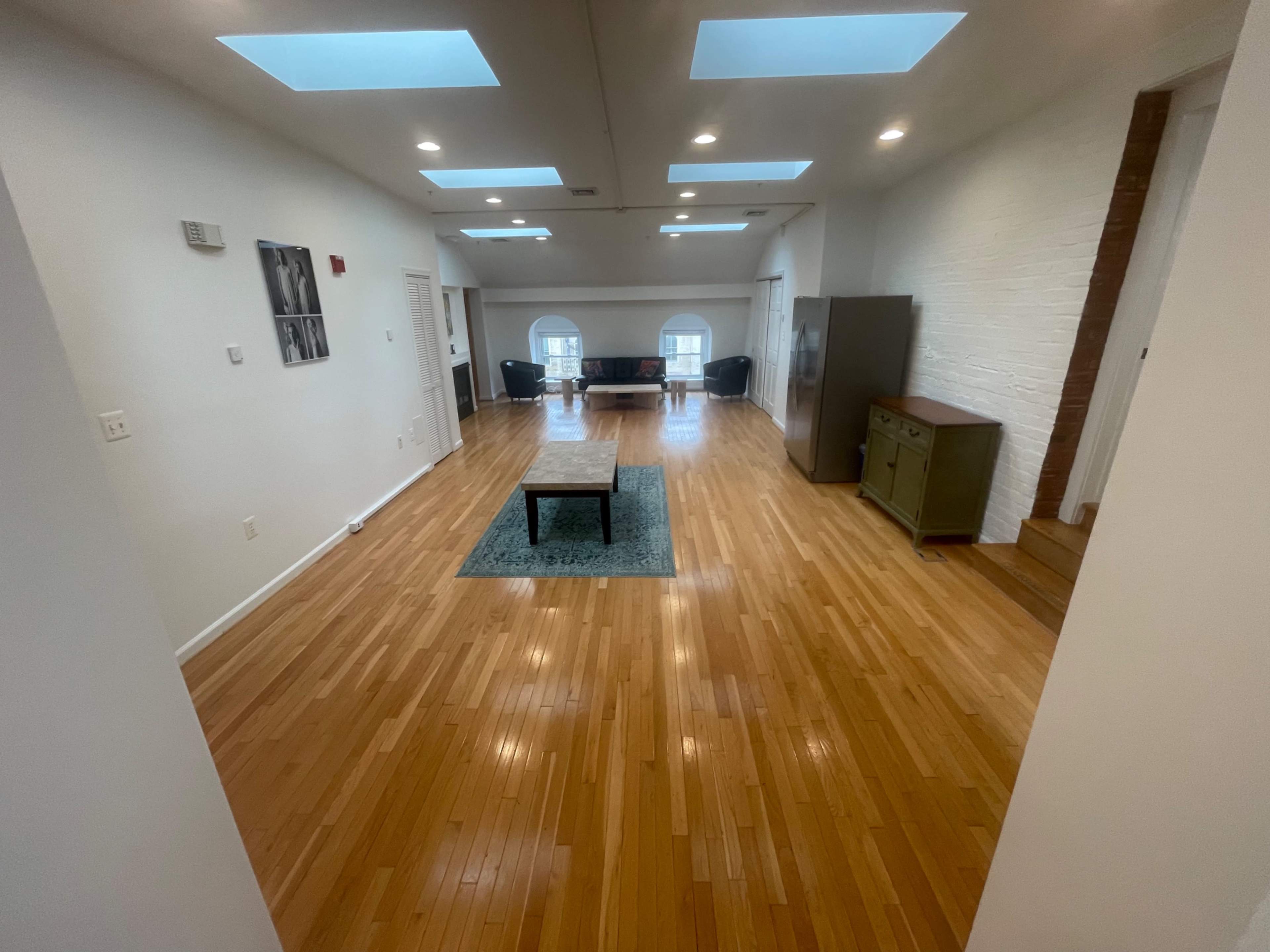 The image shows a spacious room with polished hardwood floors, large windows, and a central area featuring a small table and a decorative rug.